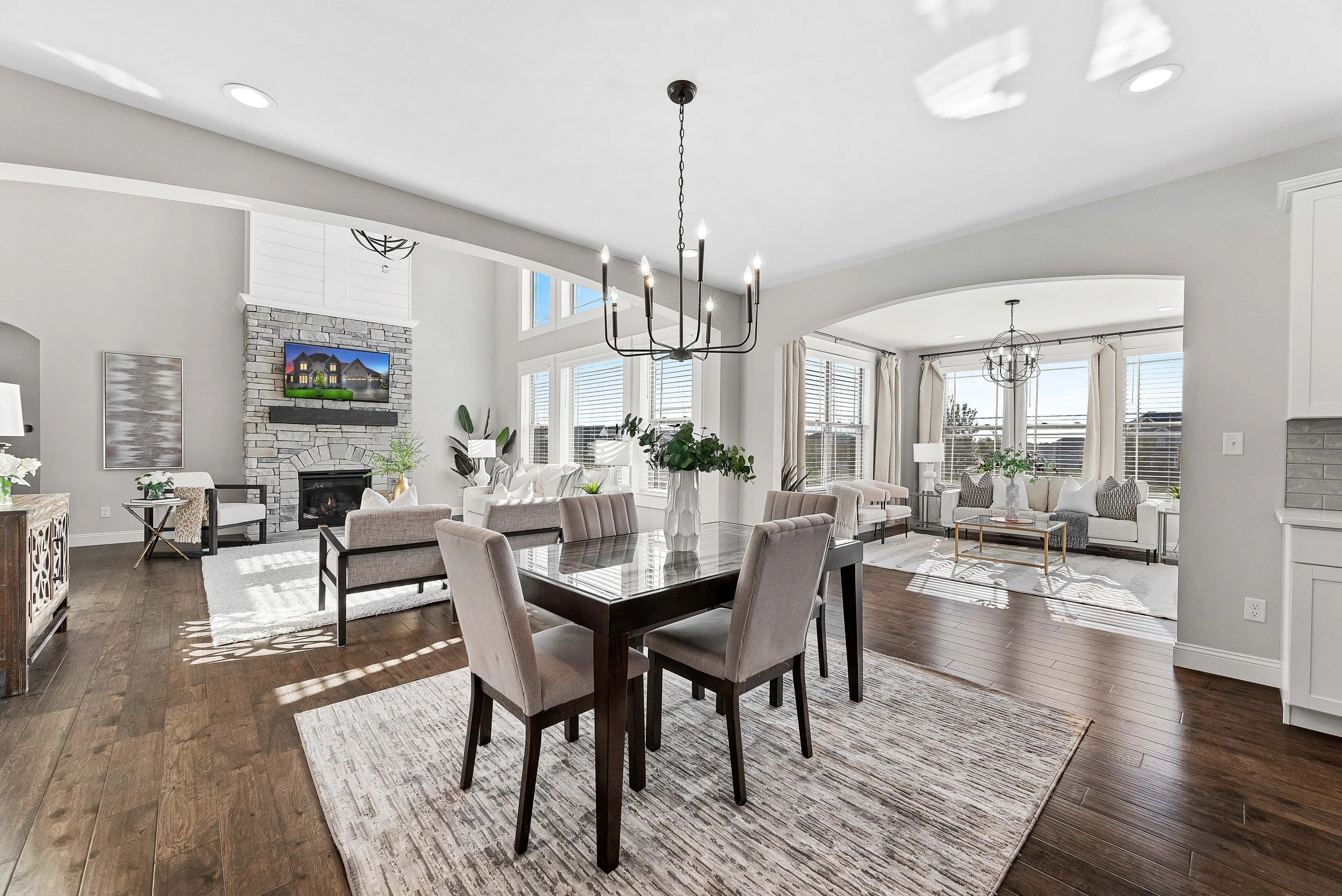Open-concept living and dining room in a modern home with white walls, large windows, hardwood floors, a stone fireplace, a dining table with six chairs, and multiple seating areas with sofas and armchairs.