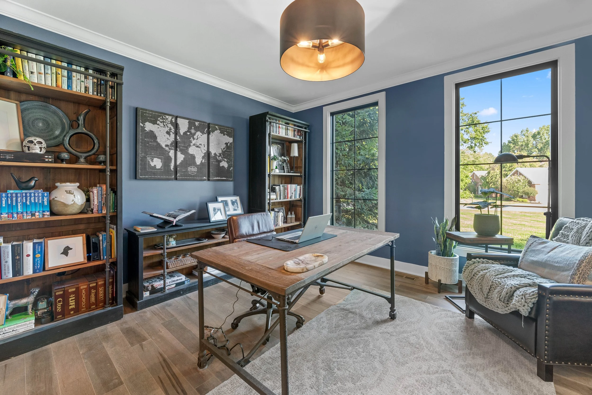 Home office with blue walls, large windows, a wooden desk with a laptop, bookshelves filled with books and decorations, a black leather chair, and a cream-colored armchair with a blanket, hardwood floors, and a ceiling light fixture.