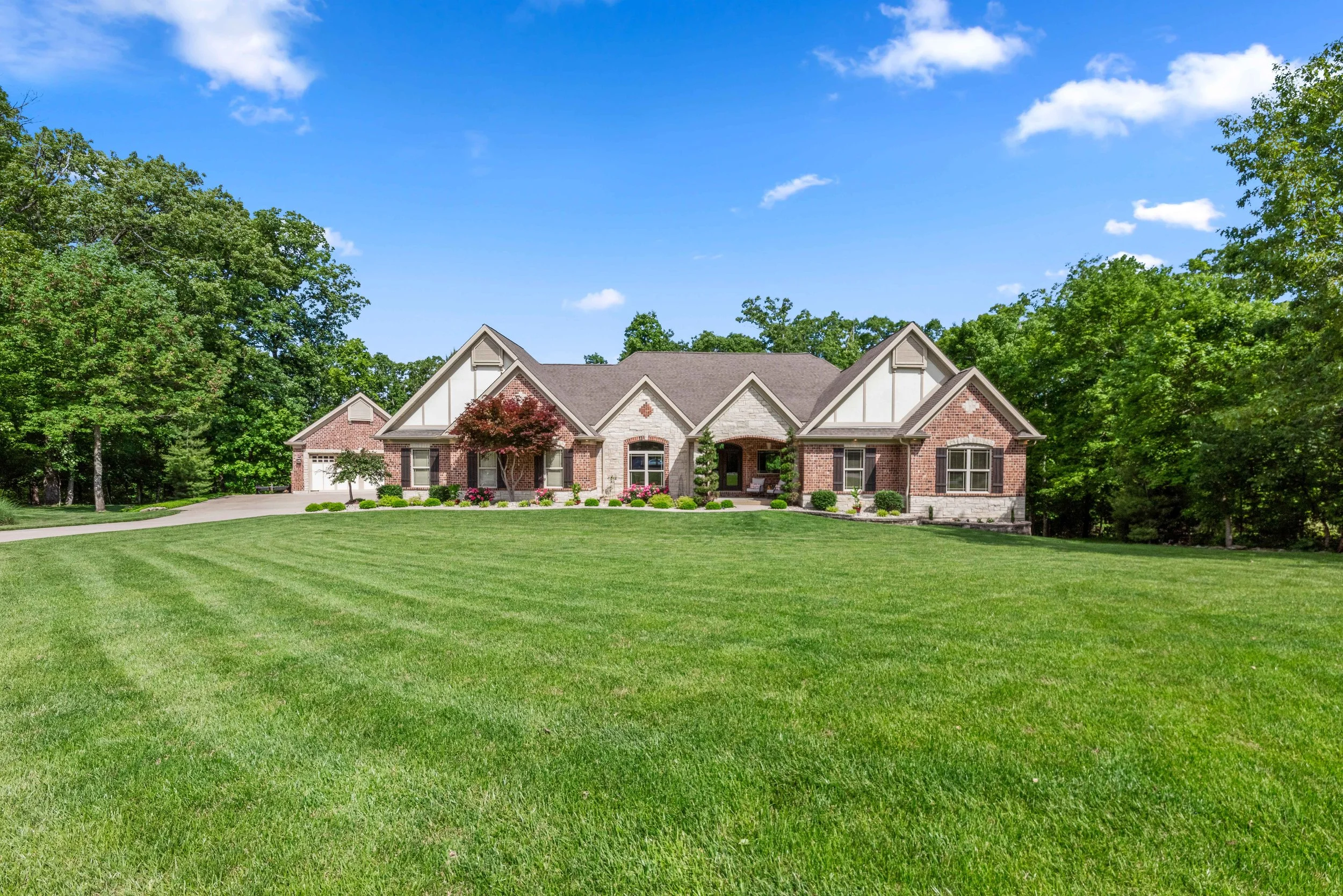 A large, modern brick house with a front porch sits on a well-maintained lawn. The house is surrounded by trees, with a clear blue sky above.