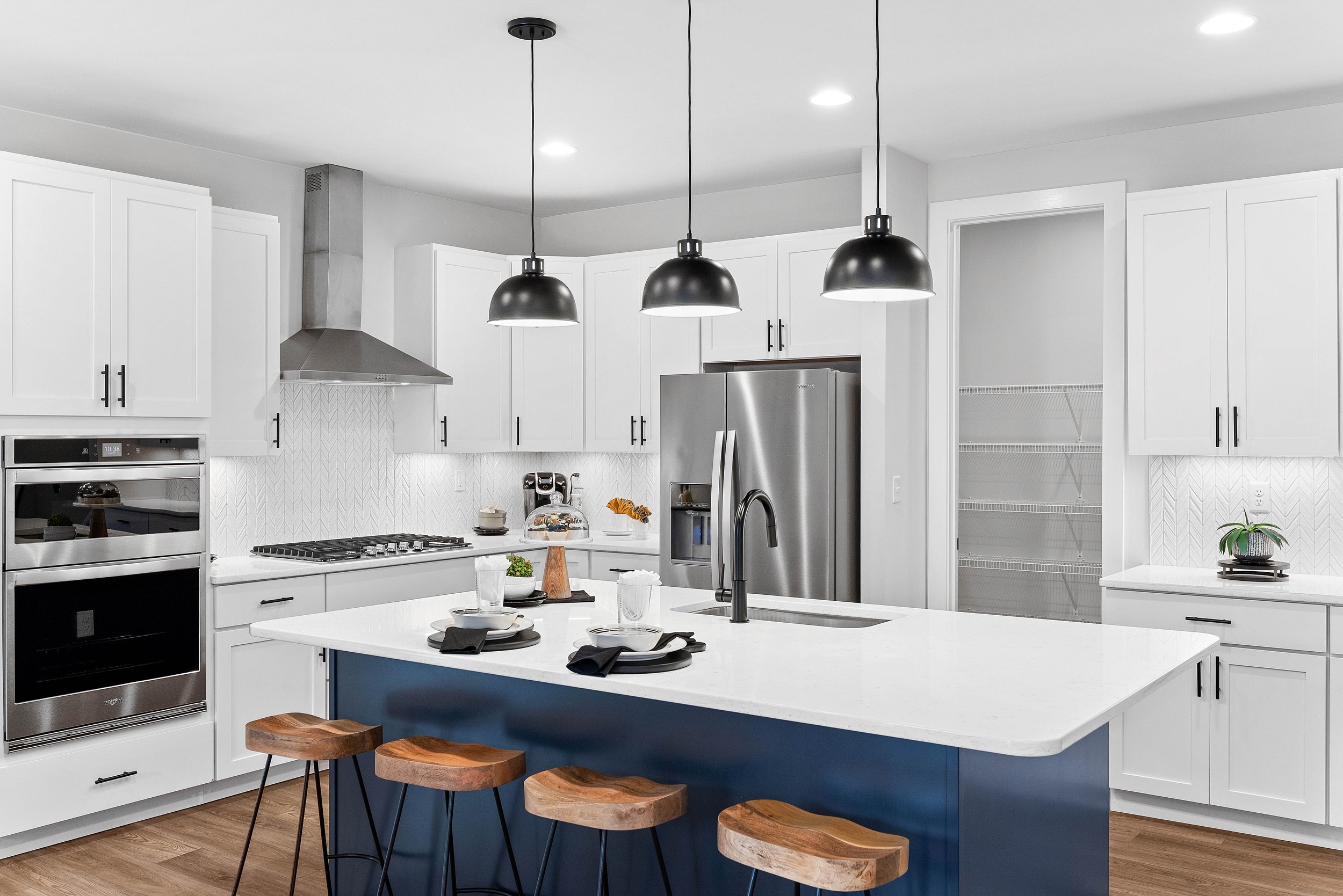 Modern kitchen with white cabinets, stainless steel appliances, black pendant lights, a blue island with wooden stools, and decorative plants.