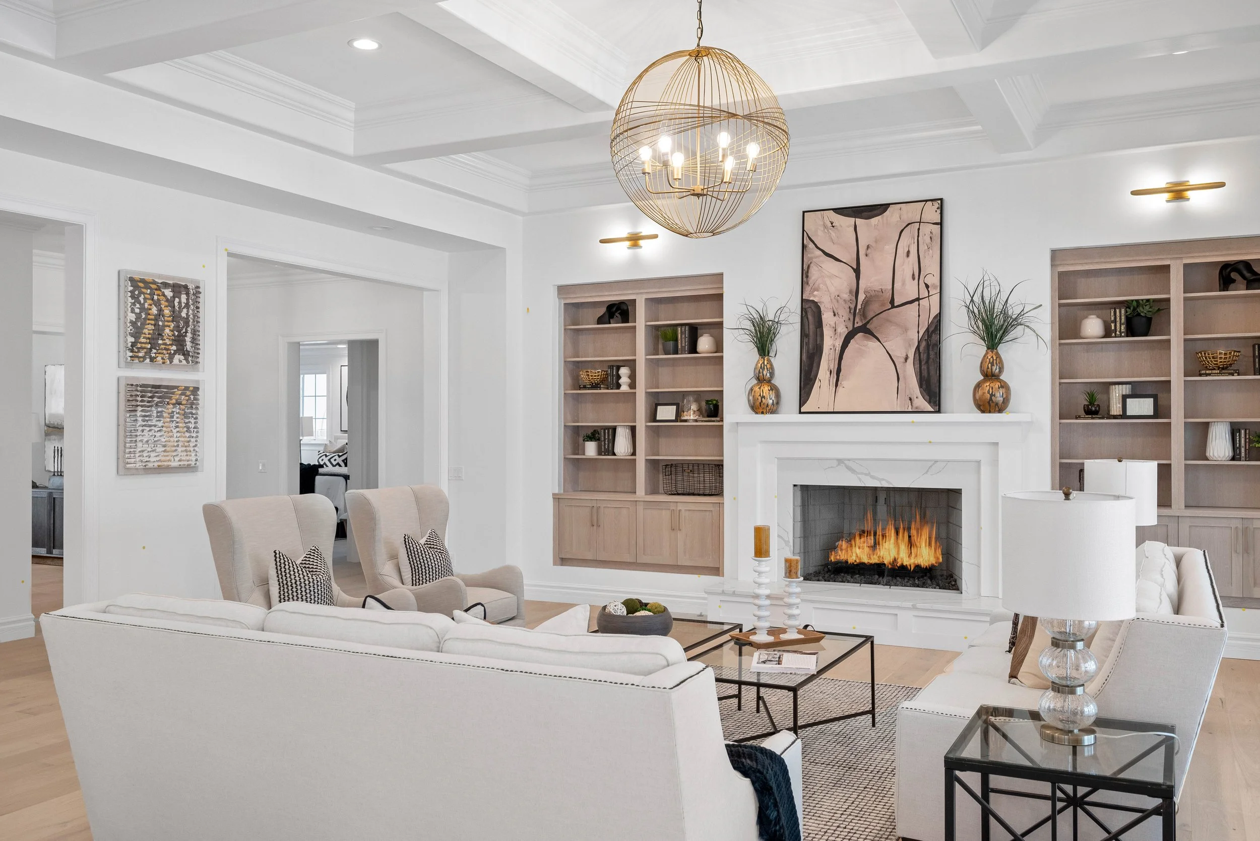 Living room with white walls, a white fireplace with a fire, built-in shelves, a large abstract painting, and a gold spherical chandelier.
