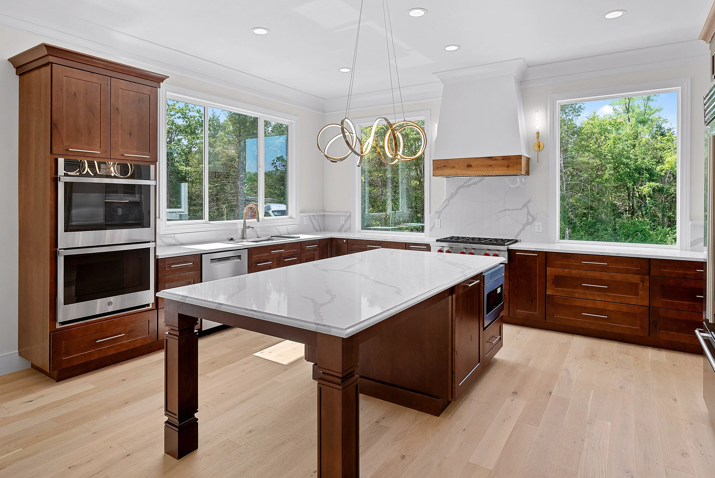 Modern kitchen with wooden cabinets, large windows, a white marble island, and stainless steel appliances.