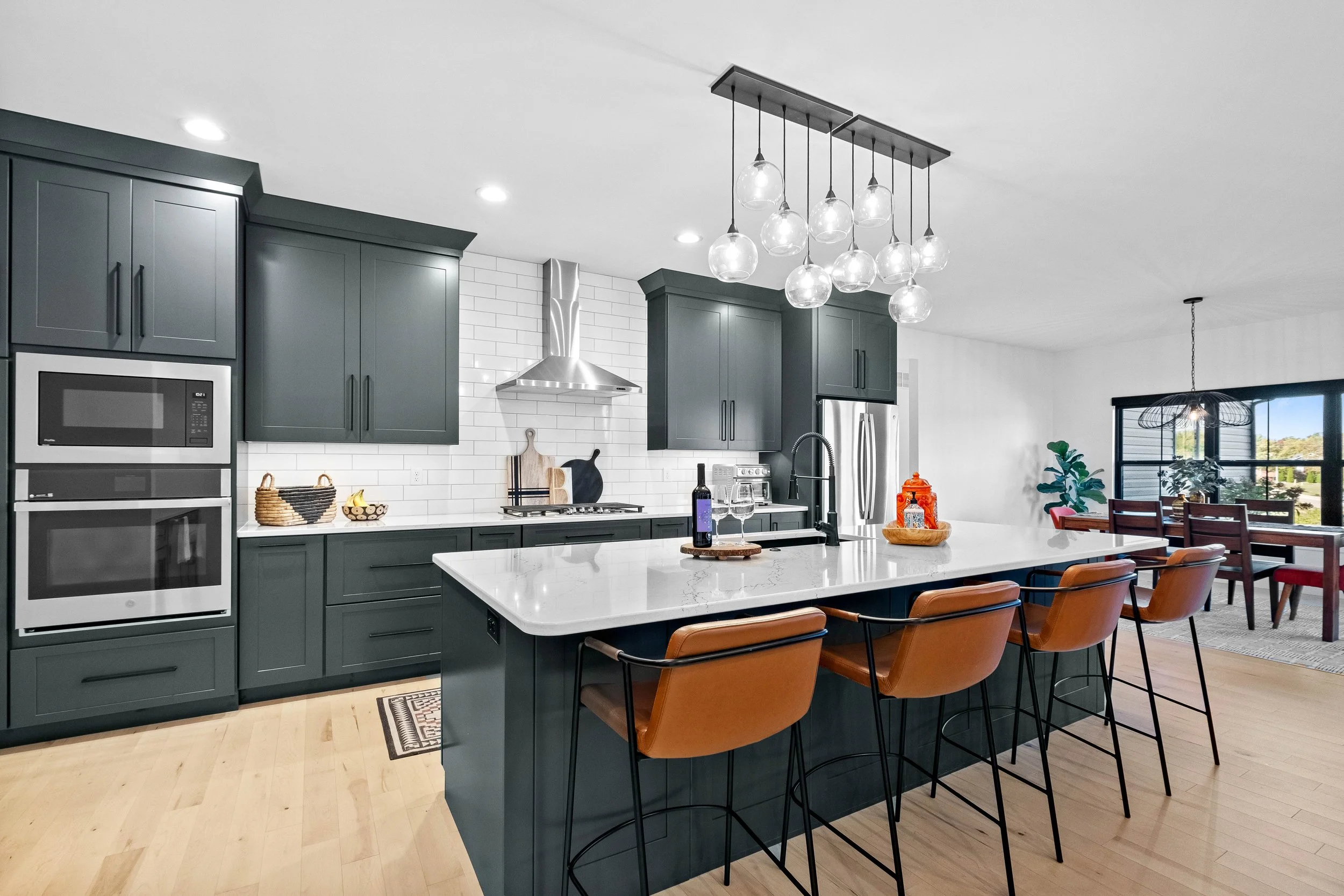 Modern kitchen with dark green cabinets, white subway tile backsplash, stainless steel appliances, a large white marble island with orange-brown barstools, a unique chandelier, and a dining area with large windows and plants.