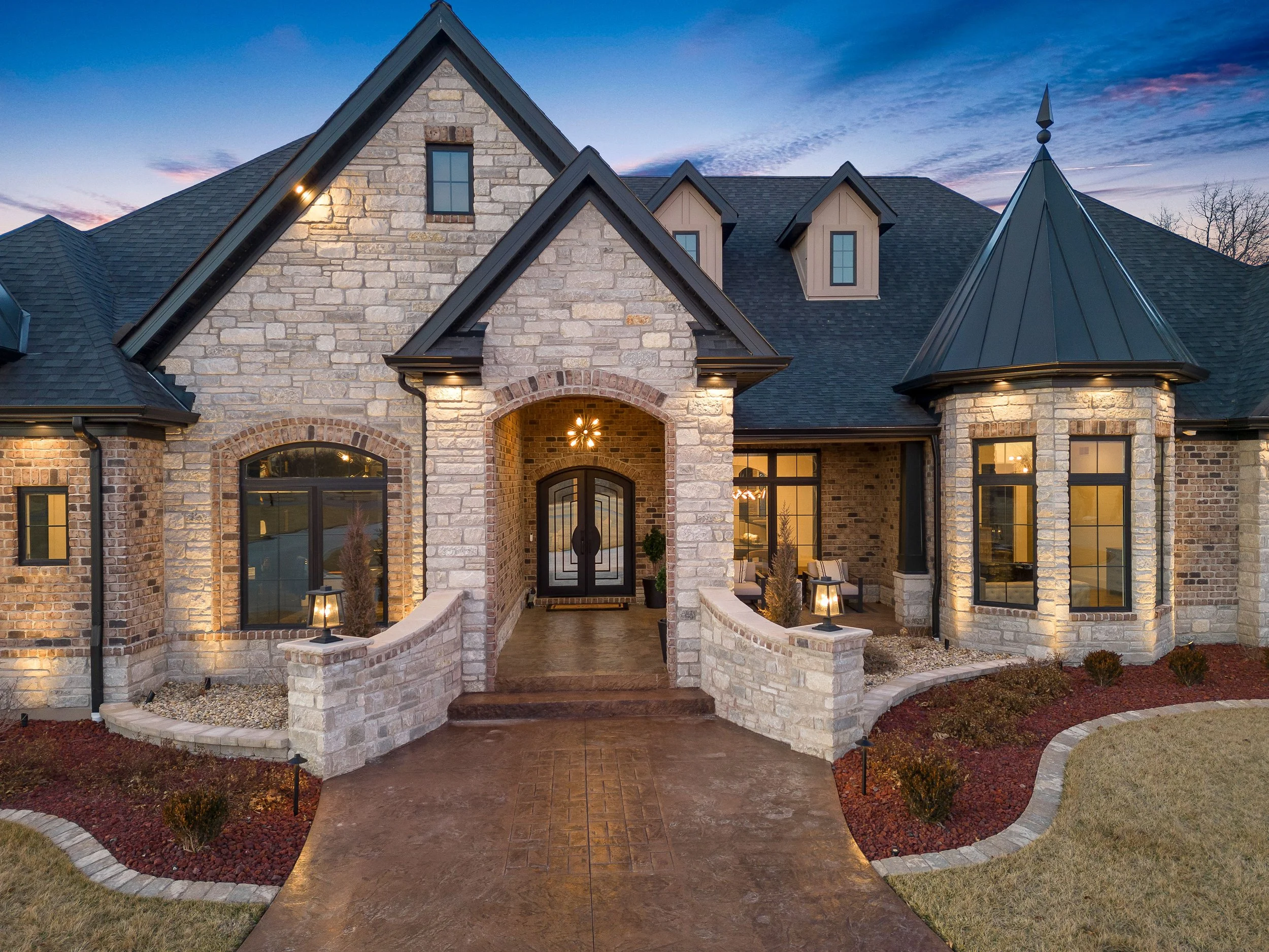 A large, elegant house with a stone facade and multiple gabled roofs during dusk, illuminated by exterior lighting, featuring a curved driveway and well-maintained landscaping.