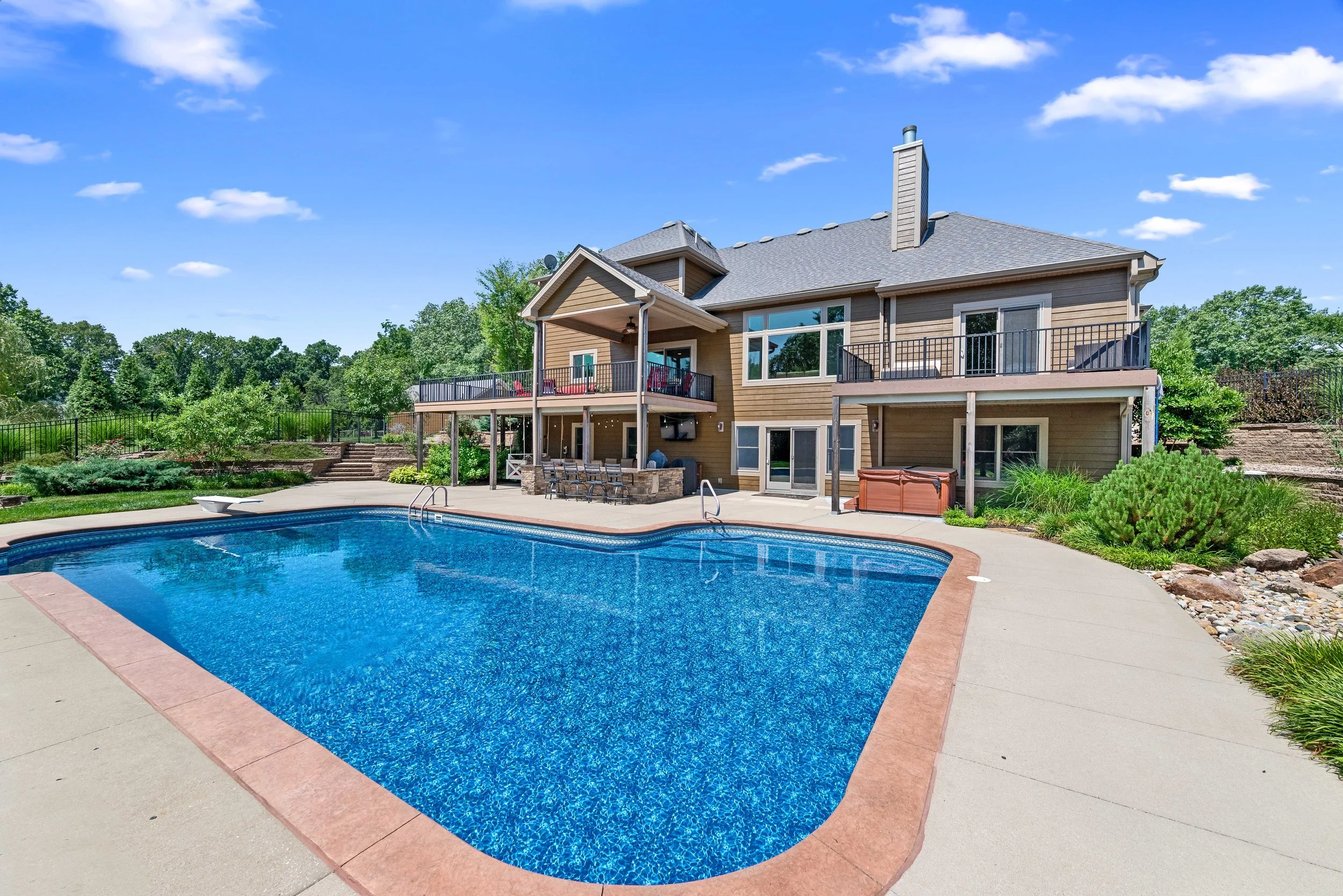 A backyard with a large swimming pool, surrounded by a concrete deck, and a two-story house with balconies and large windows in the background. The sky is blue with a few clouds, and there are green trees and landscaping around the yard.