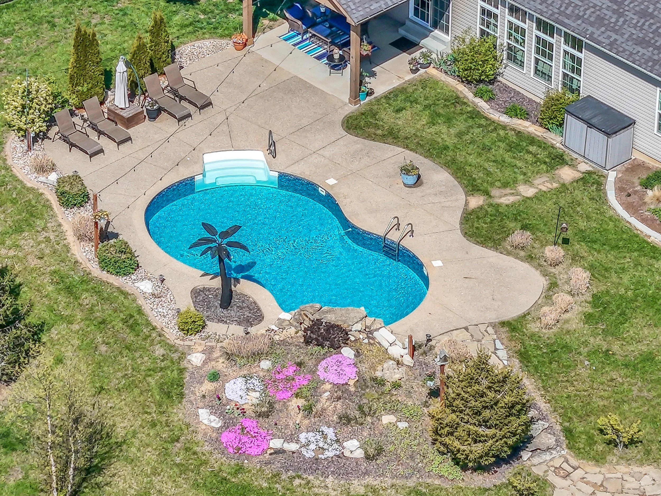A backyard with a kidney-shaped swimming pool surrounded by a concrete patio, lounge chairs, a small dog statue, and landscaped gardening with trees, bushes, and colorful flowers.
