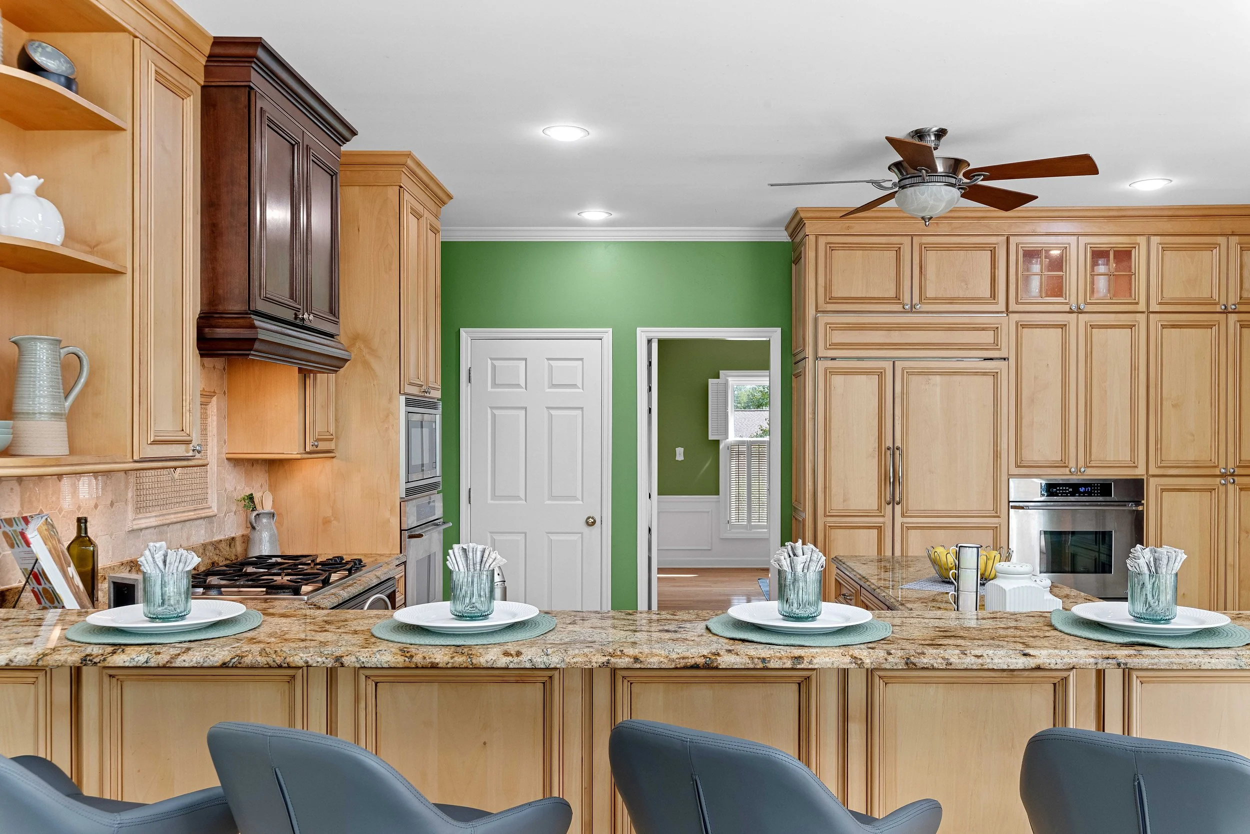 Kitchen with wooden cabinets, granite countertop, green walls, and a ceiling fan. A door leads to a room with a window, and the counter is set with four place settings, including plates, glasses, and napkins.