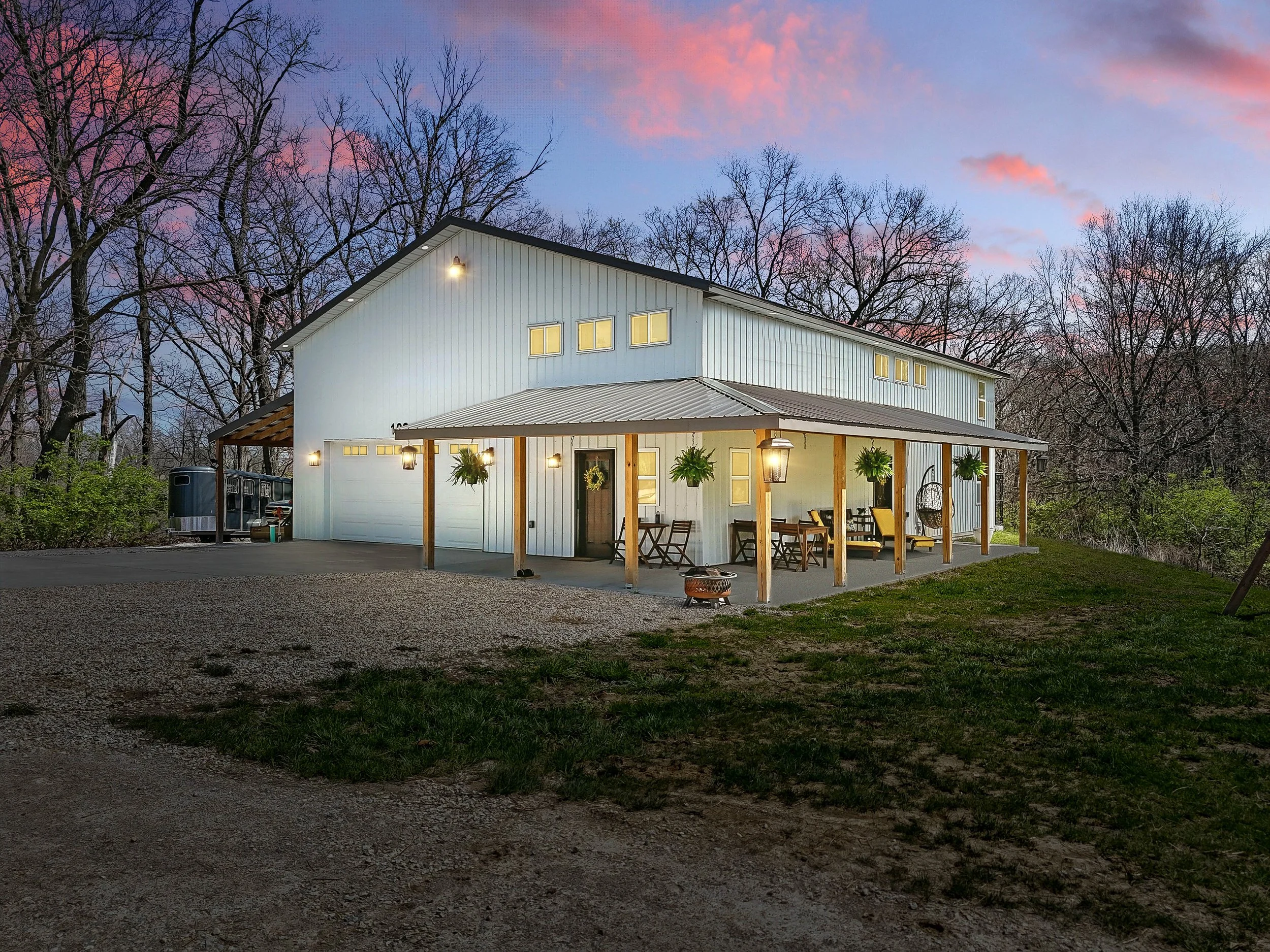 A modern farmhouse-style home with a large covered porch, outdoor seating and hanging plants, set against a backdrop of leafless trees at dusk with a colorful sky.