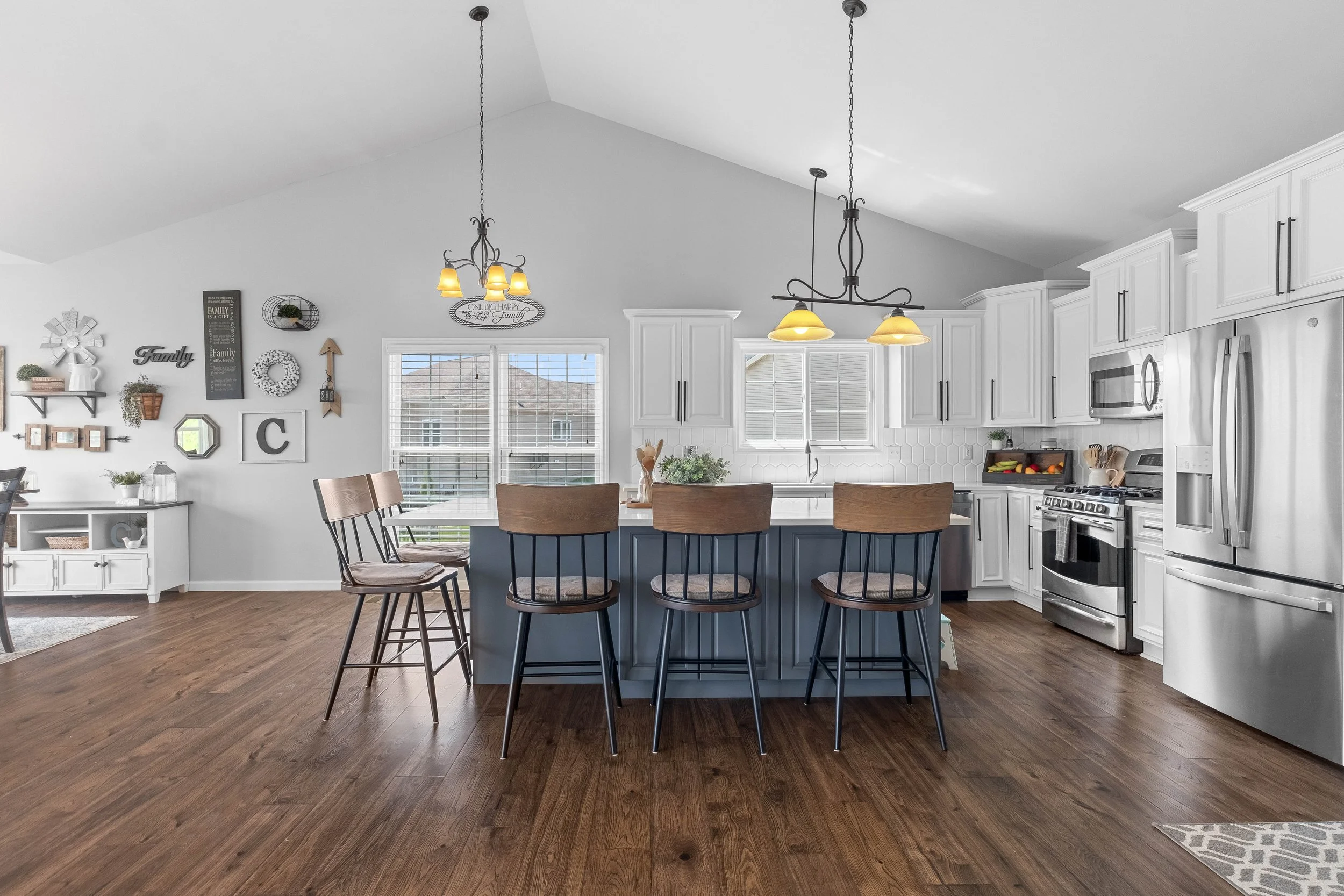 Modern kitchen with white cabinets, stainless steel appliances, a kitchen island with four wooden chairs, and a wall decorated with framed pictures and decor.