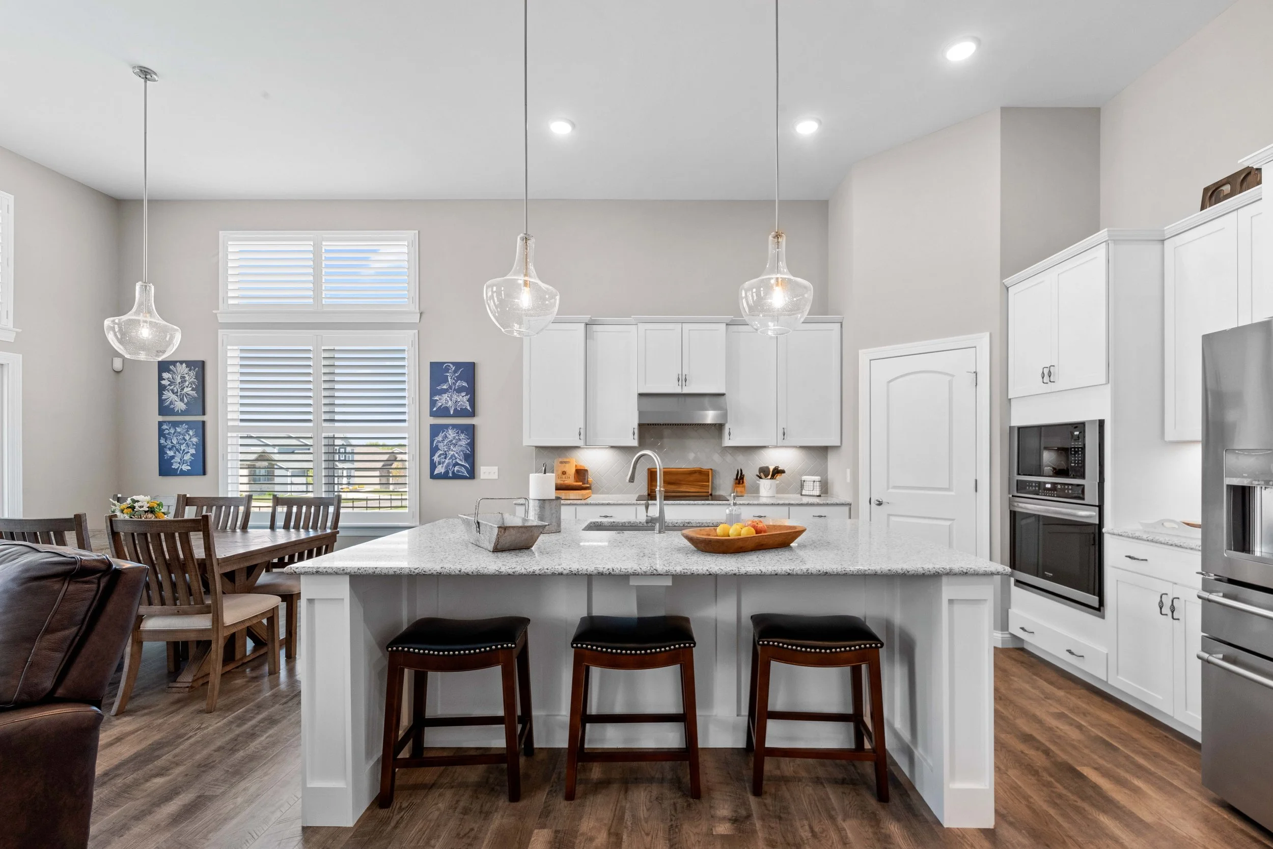 Modern kitchen with white cabinets, gray countertop island, three black stools, stainless steel appliances, and a dining area with wooden table and chairs, illuminated by hanging pendant lights.