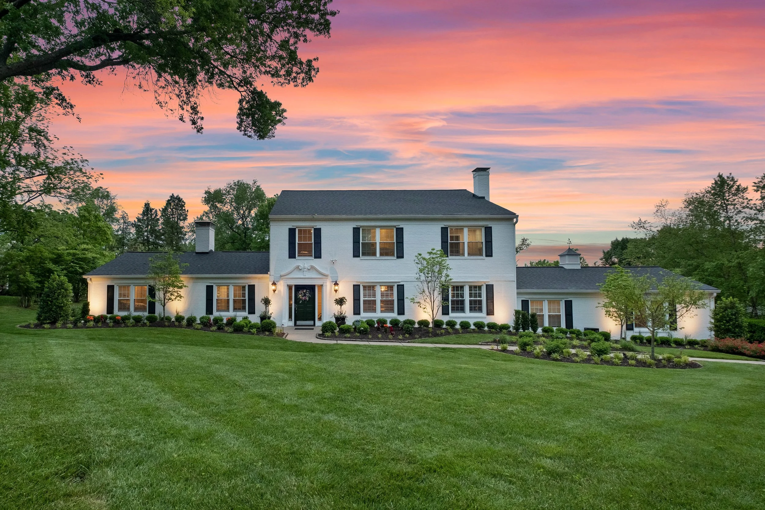 A large two-story white house with black shutters surrounded by a well-maintained lawn and landscaping under a vibrant sunset sky.