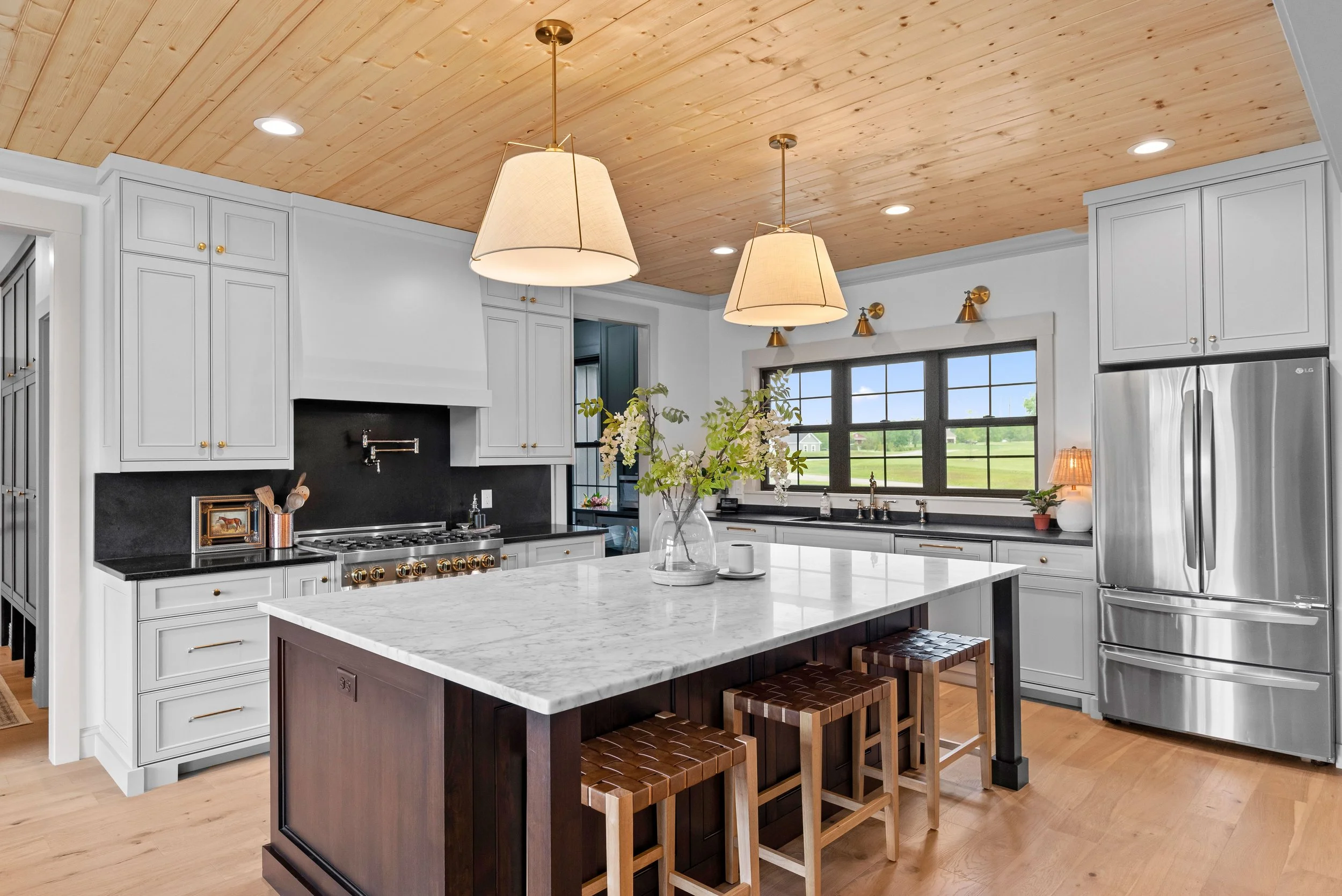 Modern kitchen with white cabinets, black countertops, stainless steel refrigerator, and wooden ceiling. An island with a white marble top and three wooden stools. Hanging pendant lights and a large window overlooking the outdoors.