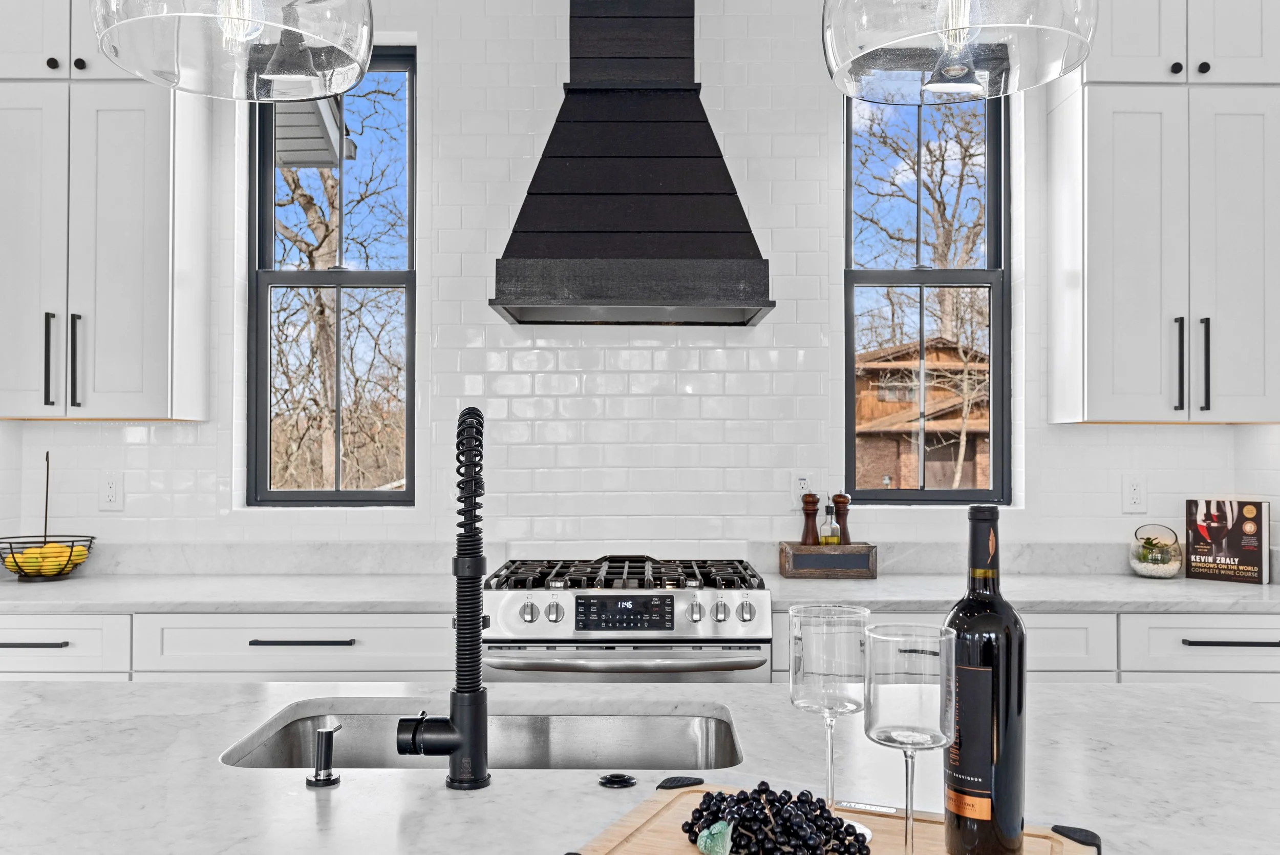 Modern kitchen with white cabinets, black kitchen hood, two windows showing outdoor trees, black faucet, wine bottle, two empty wine glasses, bunch of grapes, and a cutting board on the countertop.