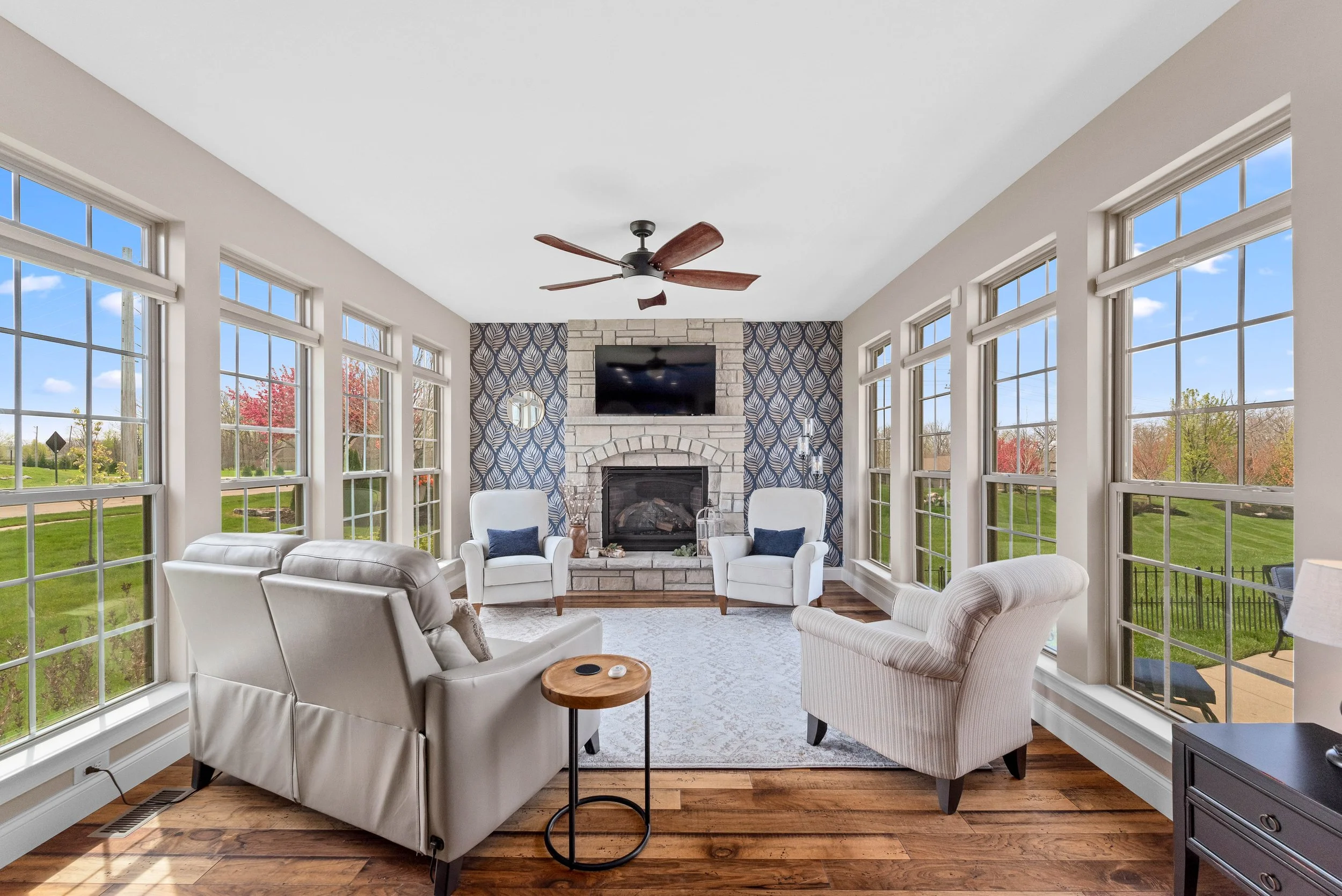 Sunlit living room with large windows, white furniture, a stone fireplace with a television above, and a view of a green yard with trees outside.