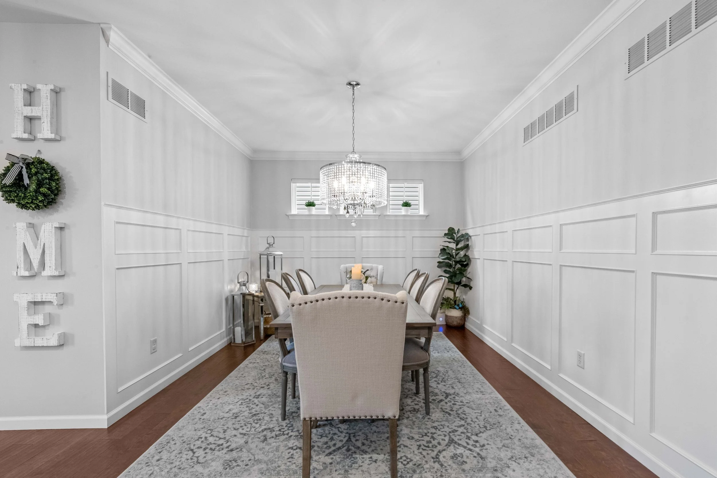 A modern dining room with a long wooden table, eight upholstered chairs, a chandelier, potted plants, and decorative wall hanging spelling "HOME".