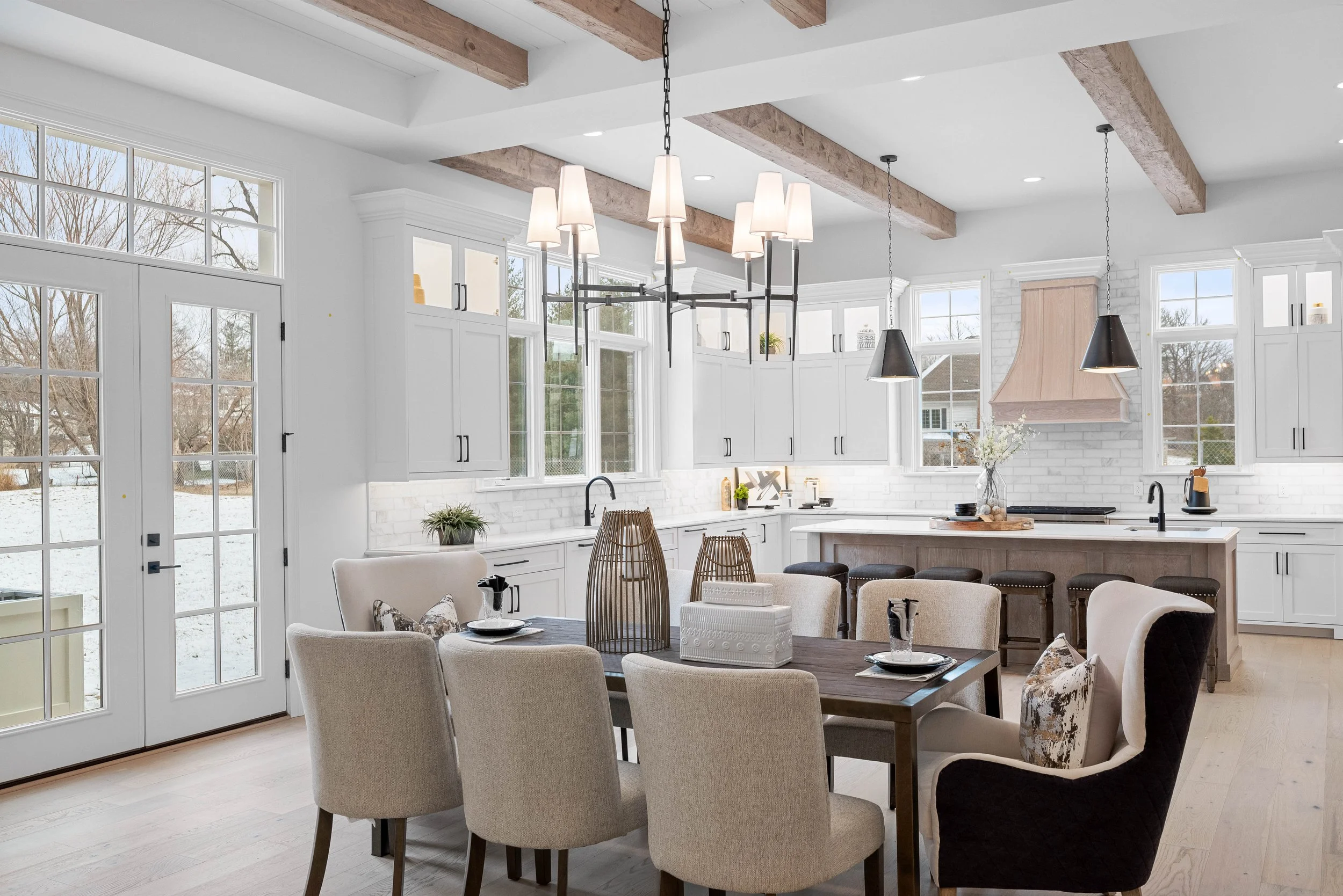 Open-concept kitchen and dining area with white cabinets, large windows, and a wooden dining table with eight beige and black upholstered chairs. Pendant lights hang above the table, and a chandelier is above the kitchen island. The room has exposed 