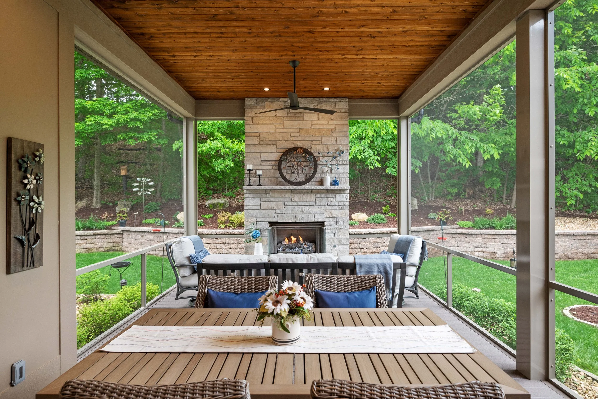 Covered porch with a wooden ceiling, a fireplace with decorative items, a dining table with a floral centerpiece, and outdoor seating facing a landscaped backyard with trees and greenery.