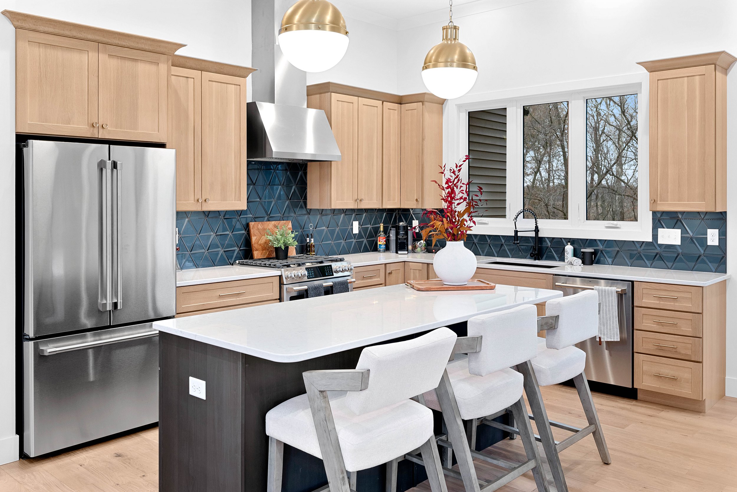 Modern kitchen with light wood cabinets, black geometric backsplash, stainless steel refrigerator, stove, and hood, white countertop, island with white surface, white chairs, large window with view of trees, vase with red and orange leaves on island.