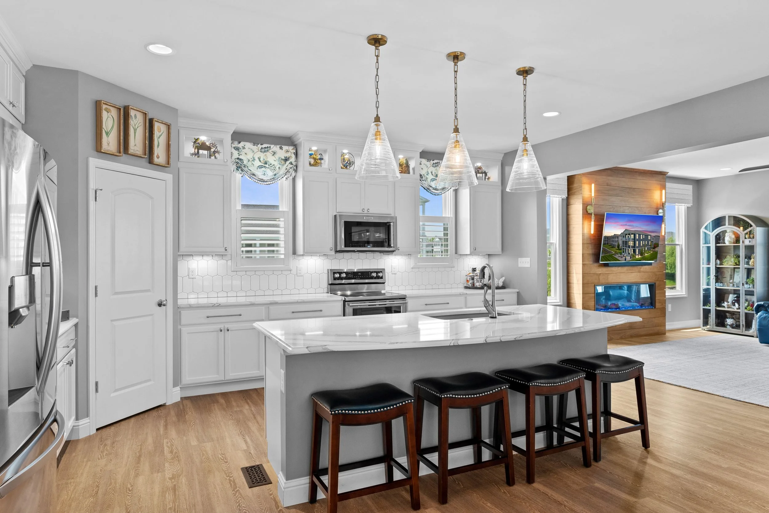 Modern kitchen with white cabinets, marble island, stainless steel appliances, three pendant lights, and a connected living area with a mounted television and a wood-paneled fireplace.