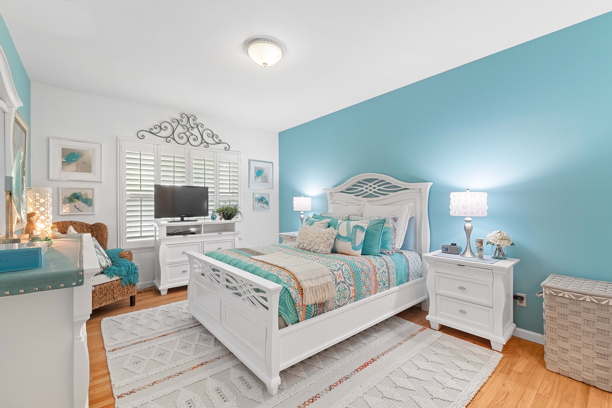 Bright bedroom with white and turquoise decor, white bed with colorful quilt and pillows, nightstand with lamp and flowers, wicker basket, and window with white shutters.