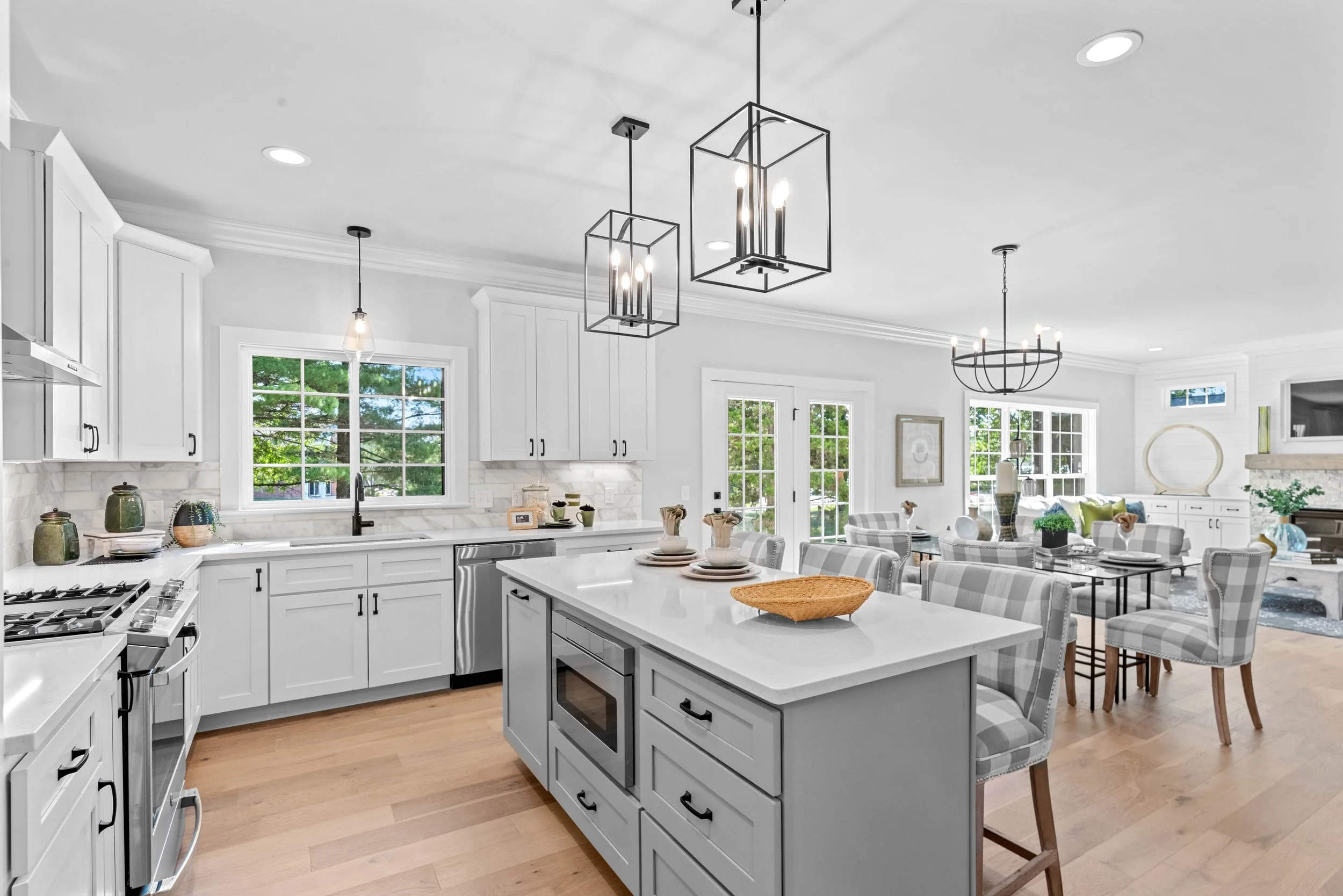 Bright open-concept kitchen and dining area with white cabinetry, a central island, light wood flooring, modern pendant lighting, and large windows and glass doors providing natural light and outdoor views.