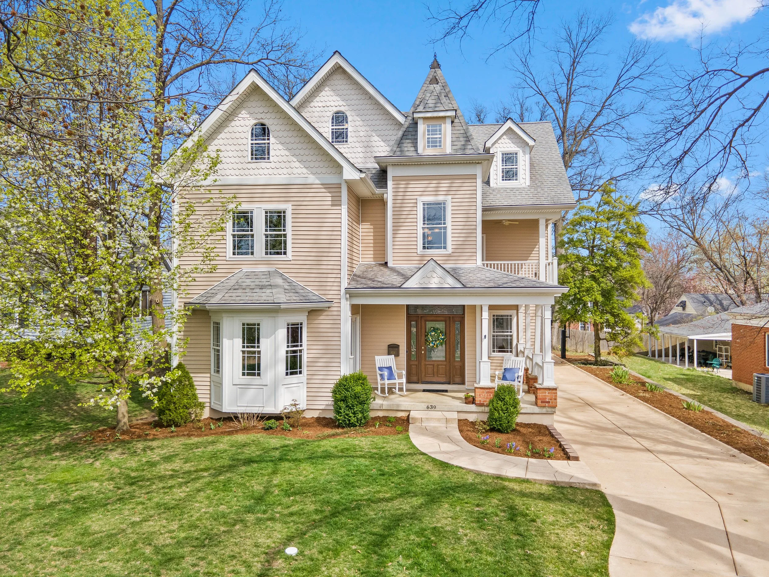 A large, beige, multi-story house with a front porch, surrounded by trees and a well-maintained lawn, under a partly cloudy blue sky.