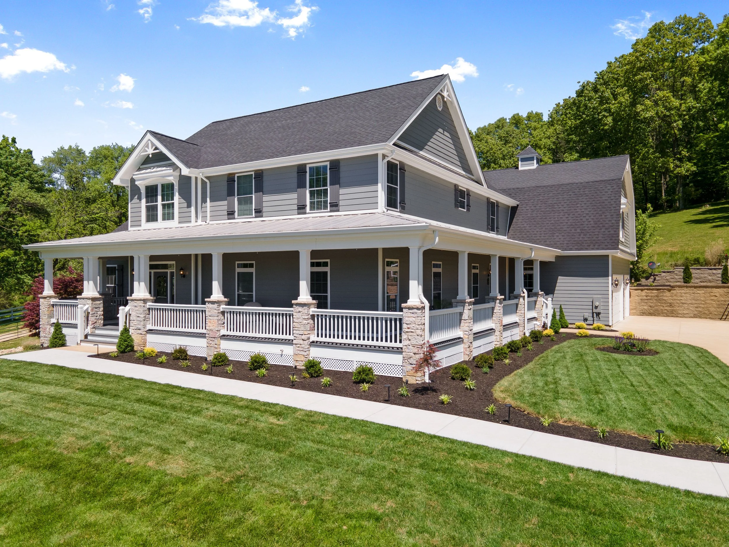A two-story house with blue siding, white trim, a covered front porch, and a well-maintained lawn with landscaping, trees in the background, and a clear blue sky.