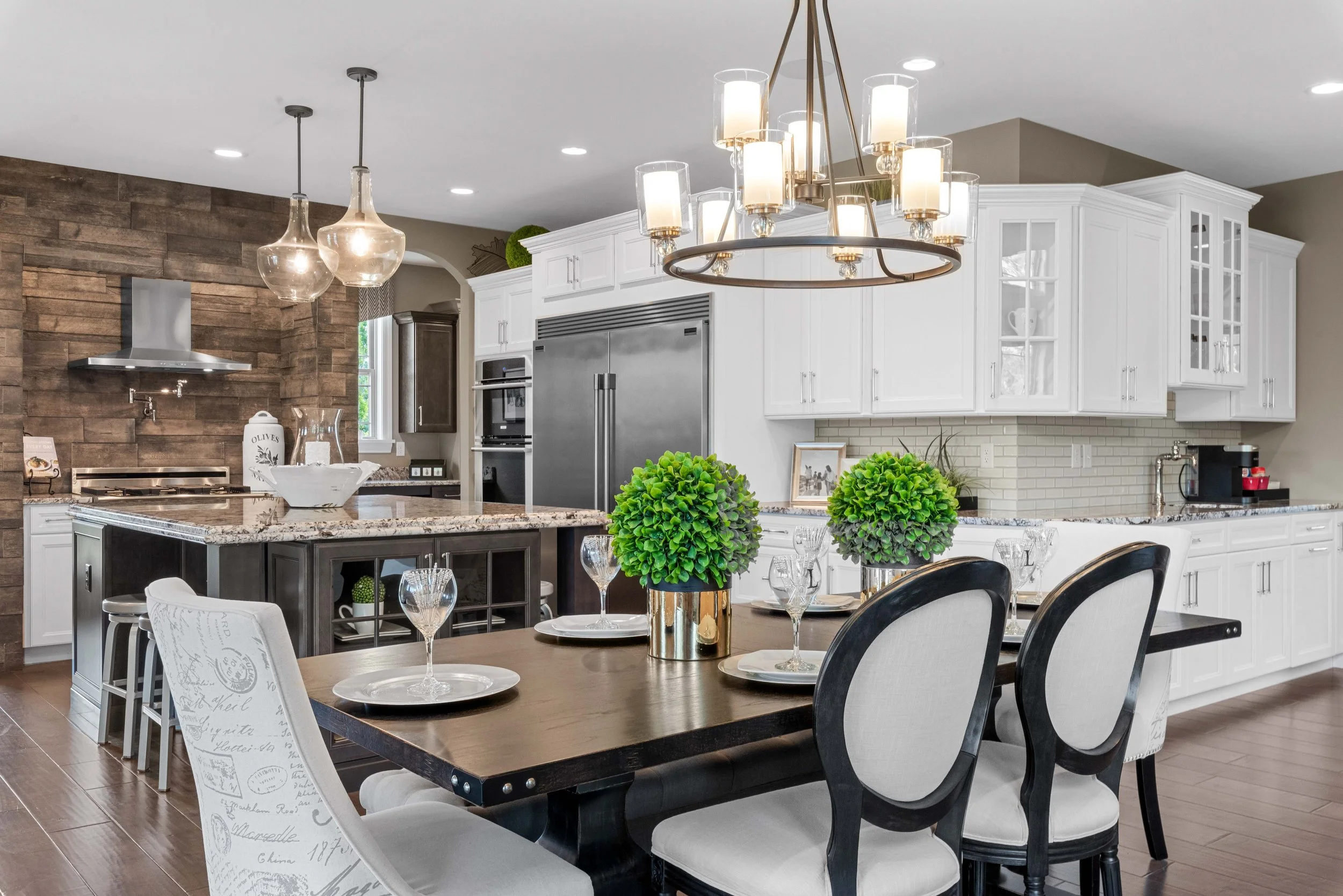 Modern kitchen with white cabinets, stainless steel appliances, a central island, a dining table with chairs and green plants, pendant and chandelier lighting, wood and brick accents.