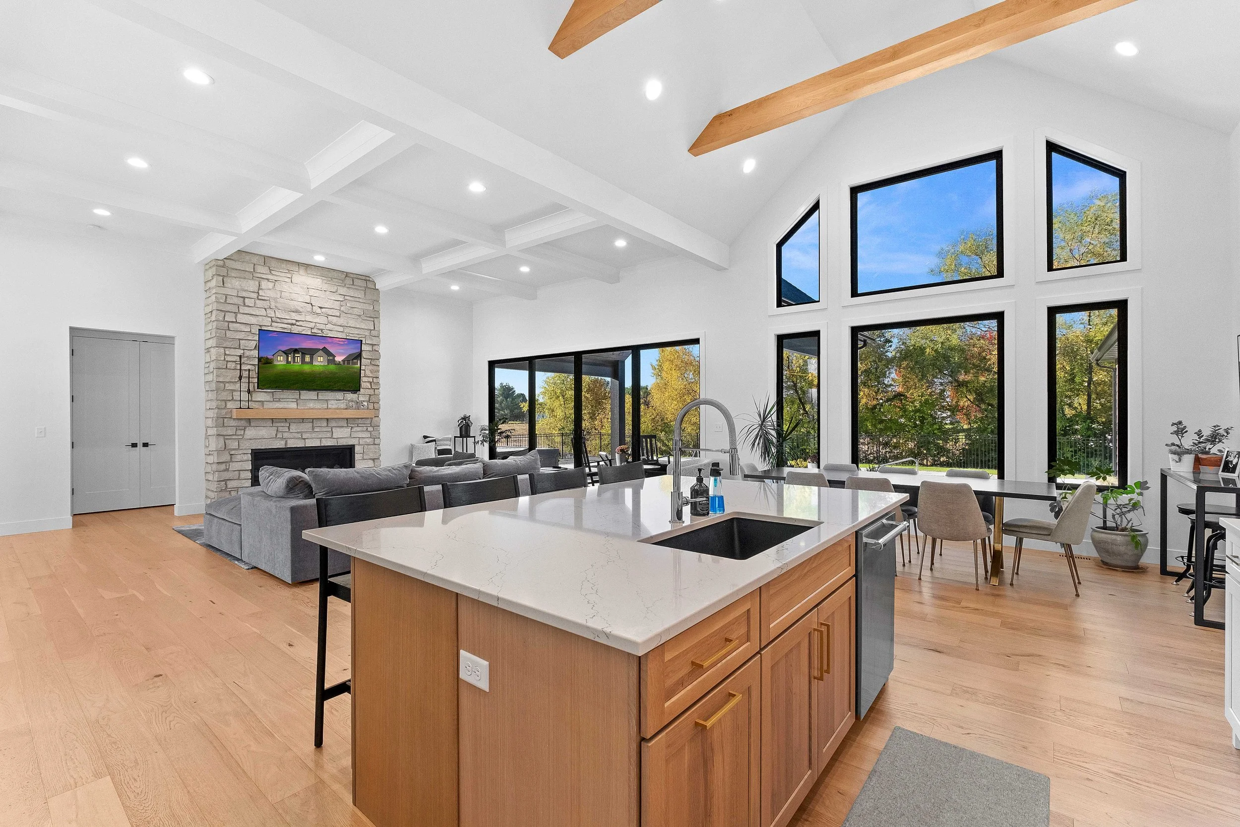 Open-concept living room and kitchen with large windows, a stone fireplace, a gray sectional sofa, and wooden ceiling beams.