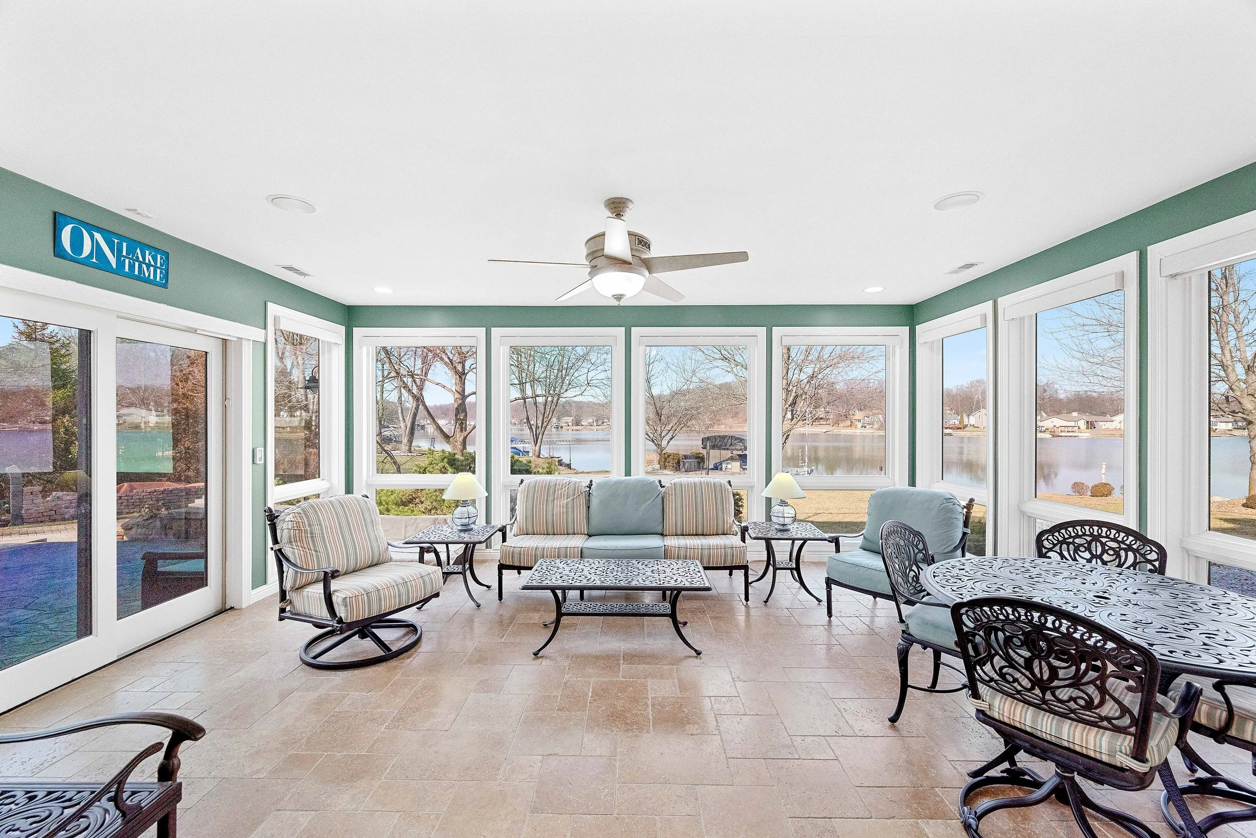 Sunroom with large windows overlooking a lake, furnished with a striped sofa, matching chairs, a metal dining table with ornate chairs, side tables with lamps, and a ceiling fan.