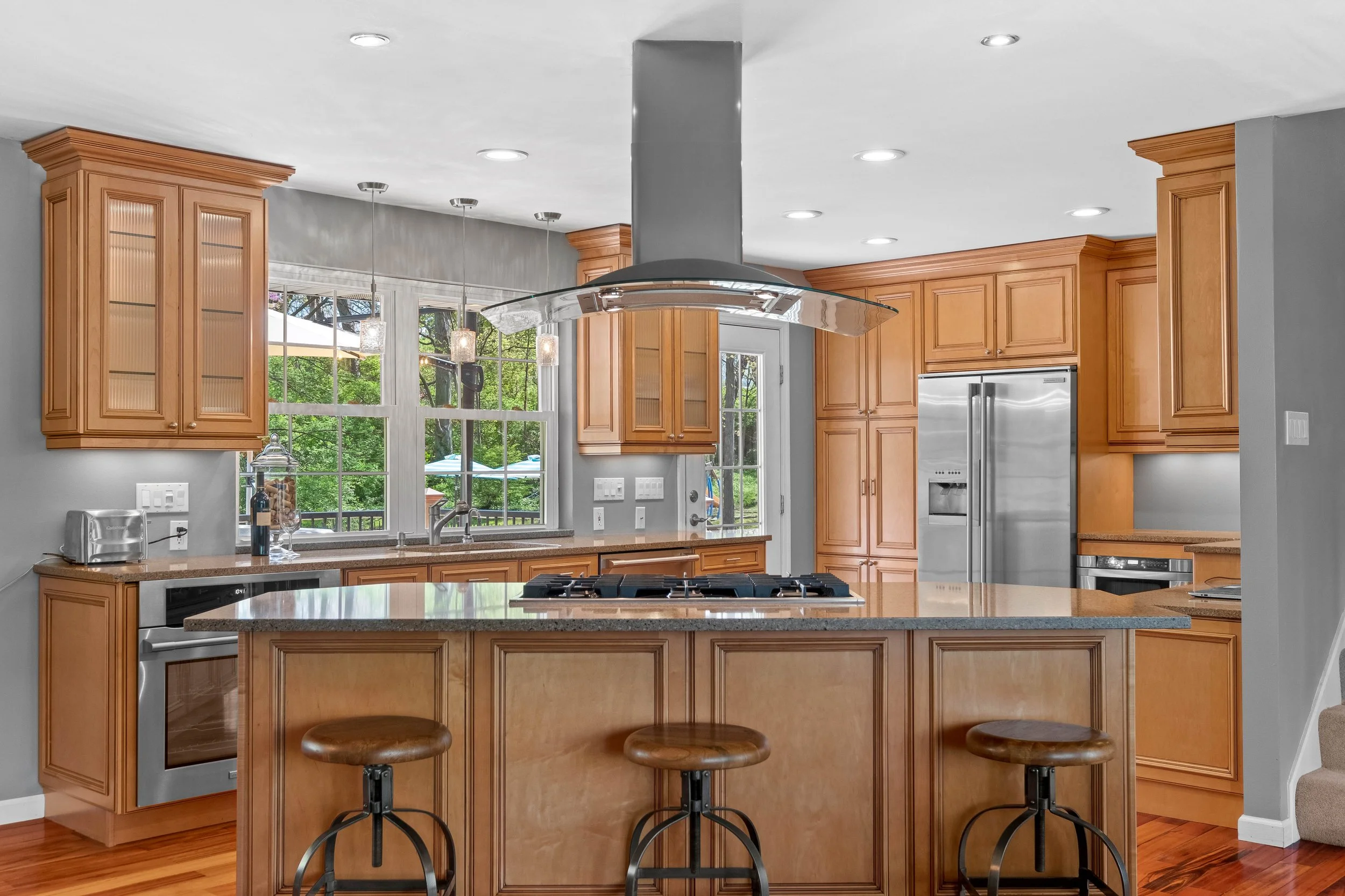 Kitchen with wooden cabinets, an island with bar stools, stainless steel appliances, and a window overlooking a backyard.