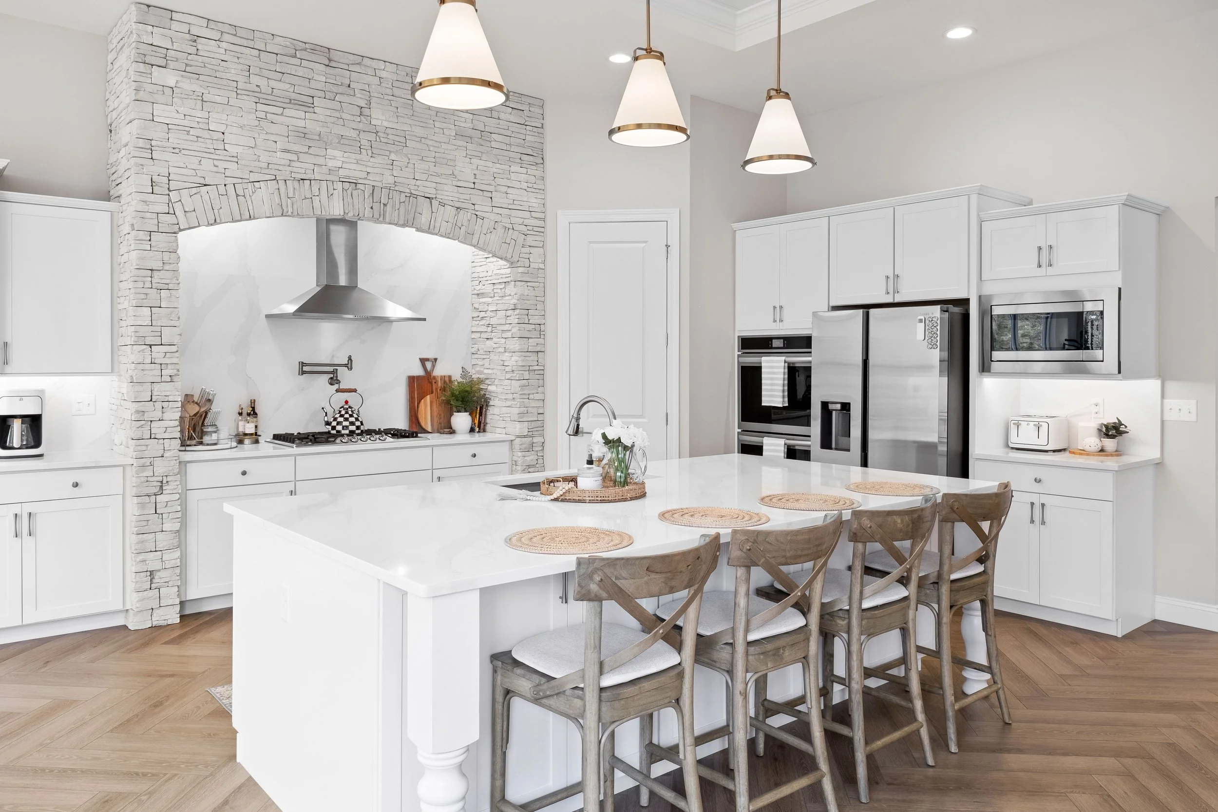 A modern kitchen with white cabinetry, a large island with a white countertop, and four wooden chairs. There is a stone arch around the stove area with a stainless steel range hood, and built-in appliances including a refrigerator, oven, and microwav