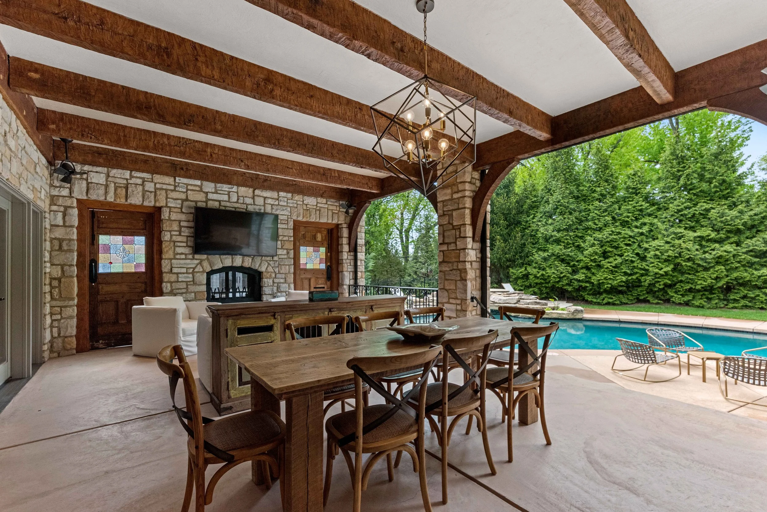 Covered outdoor patio with a wooden dining table and chairs, rustic stone wall with a fireplace and a mounted flat-screen TV, overlooking a swimming pool surrounded by lounge chairs and lush green trees.