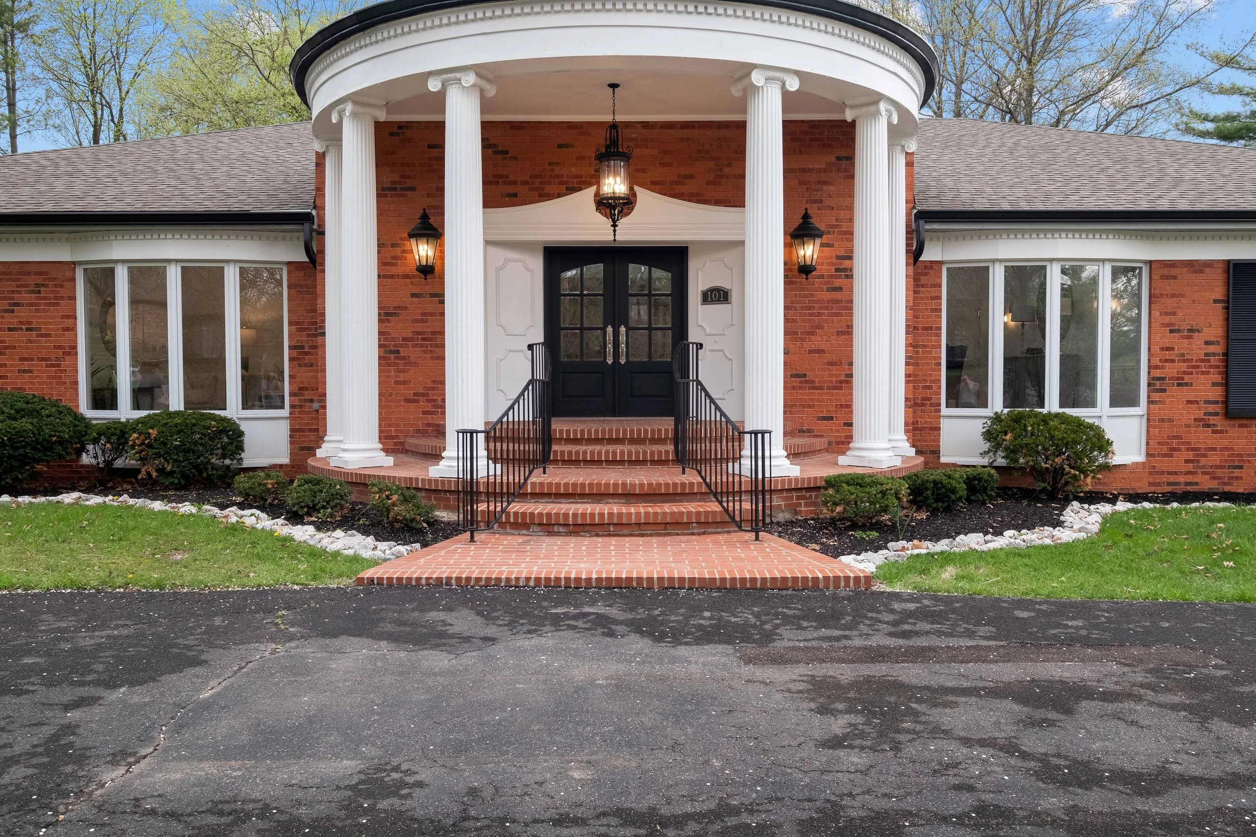 Front view of a brick house with a rounded porch supported by white pillars, black double doors, and a brick staircase with black railings leading to the entrance. The house has large windows on either side of the door, landscaped bushes, and outdoor
