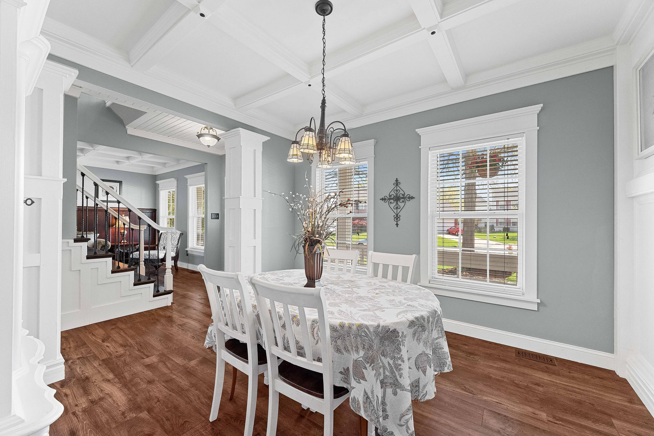 Dining room with four white chairs around a round table covered with a floral tablecloth, a brown vase with flowers on the table, large windows with white blinds, a chandelier hanging from a coffered ceiling, and wooden flooring.