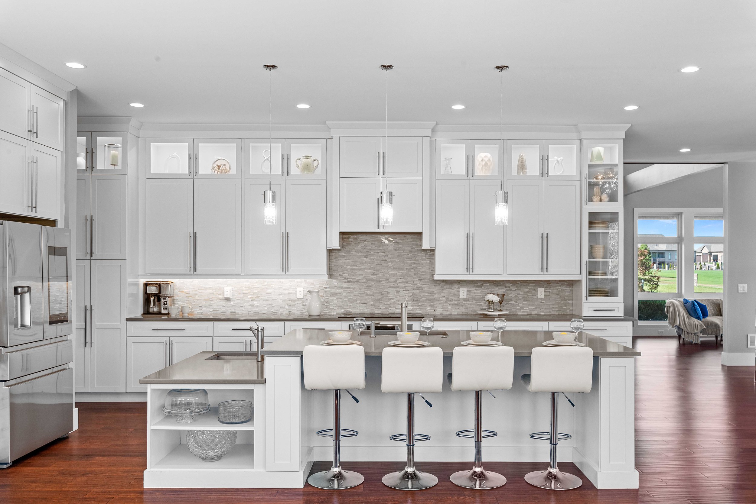 Modern white kitchen with island counter, four white barstools, pendant lighting, wood flooring, and a view into a living room with large windows