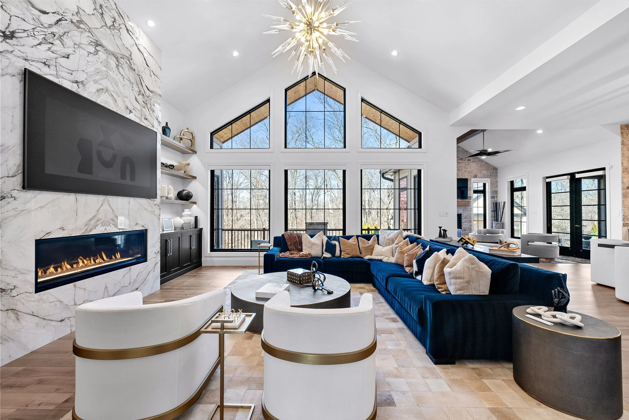Spacious living room with a high vaulted ceiling, large windows, a wall-mounted TV above a modern fireplace, a large blue sectional sofa with multiple pillows, a round coffee table, and two white armchairs, with a view of an outdoor deck through the 