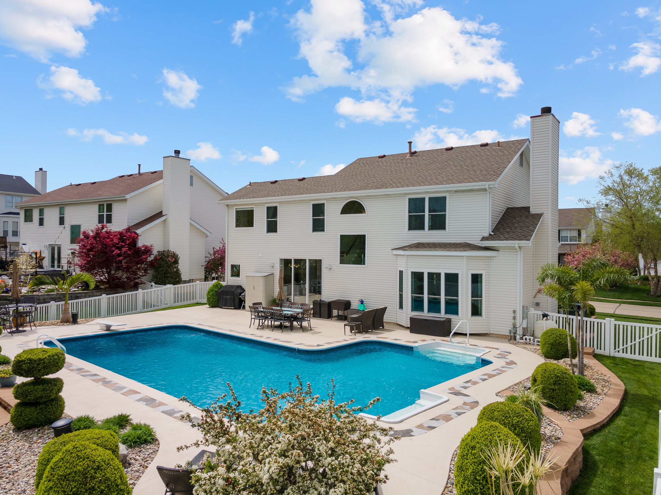 Backyard with swimming pool, patio furniture, and a white house with multiple windows under a partly cloudy sky.