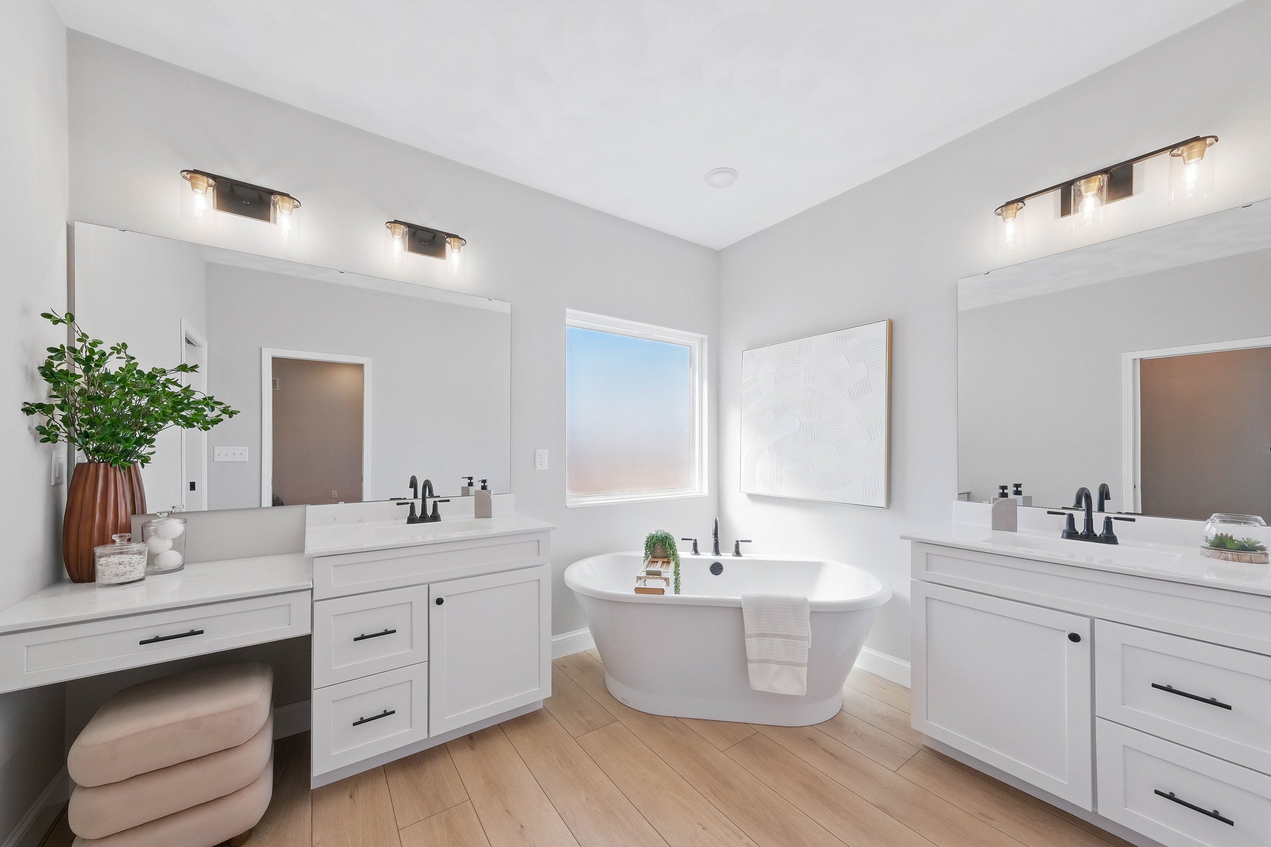 Bright modern bathroom with a white bathtub between two white vanities with black fixtures, a large mirror above each vanity, a window, and a large white abstract painting on the wall.