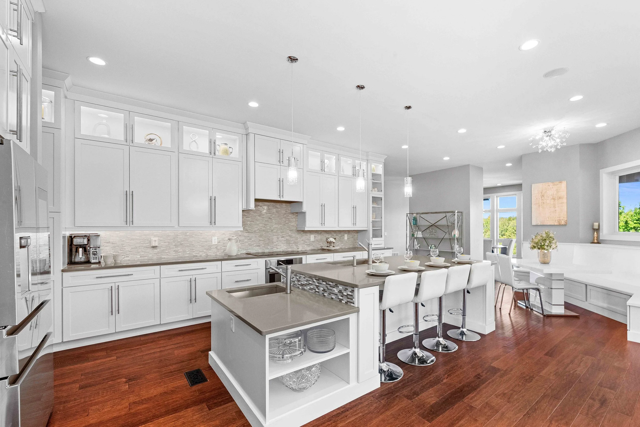 Bright modern kitchen with white cabinets, a spacious island with a beige countertop, and hardwood floors. There are four white barstools at the island, pendant lights hanging above, and a dining area with a window view outside.