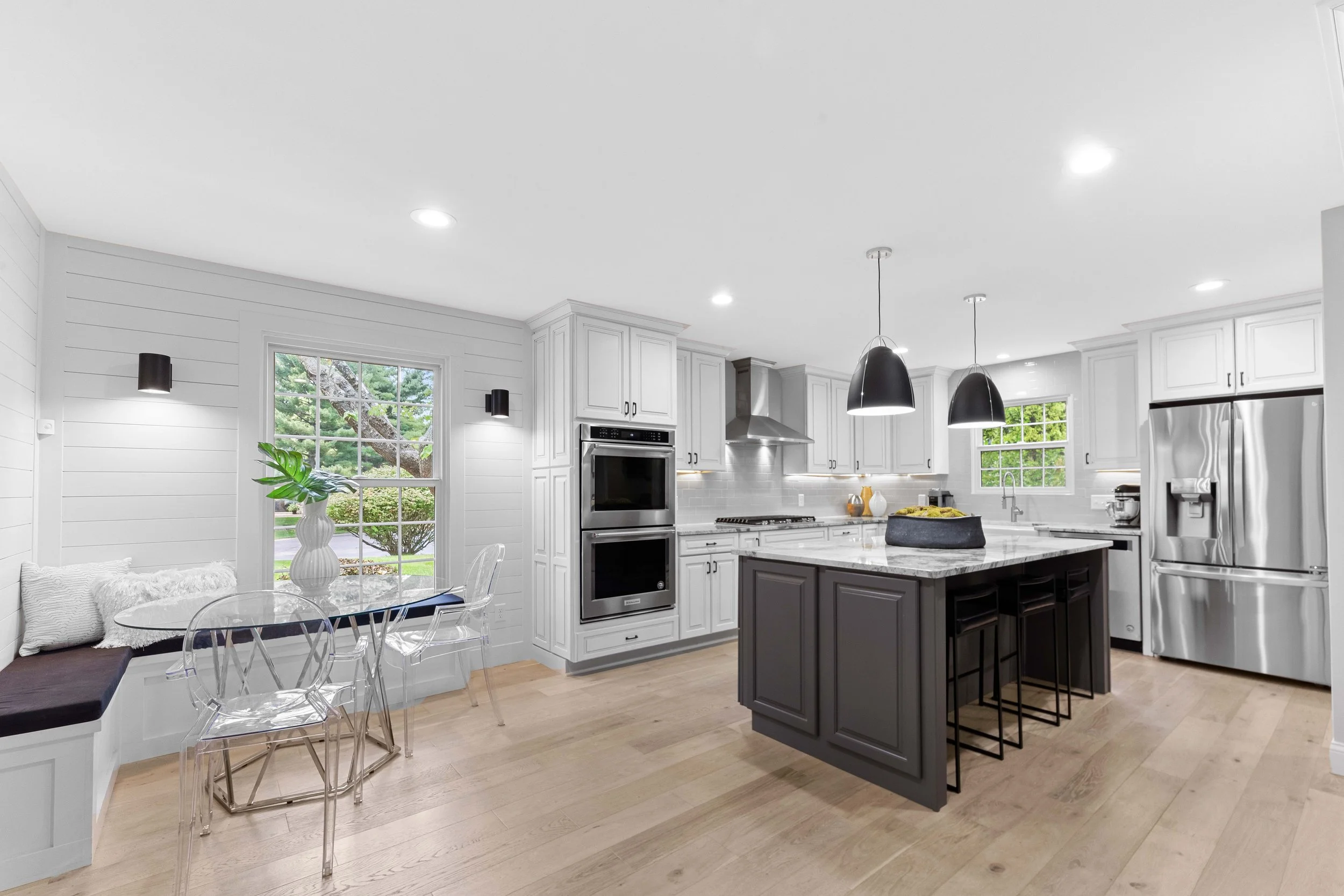 Modern white kitchen with black island and stainless steel appliances, including refrigerator and double oven, with a small breakfast nook by the window and black pendant lights hanging over the island.