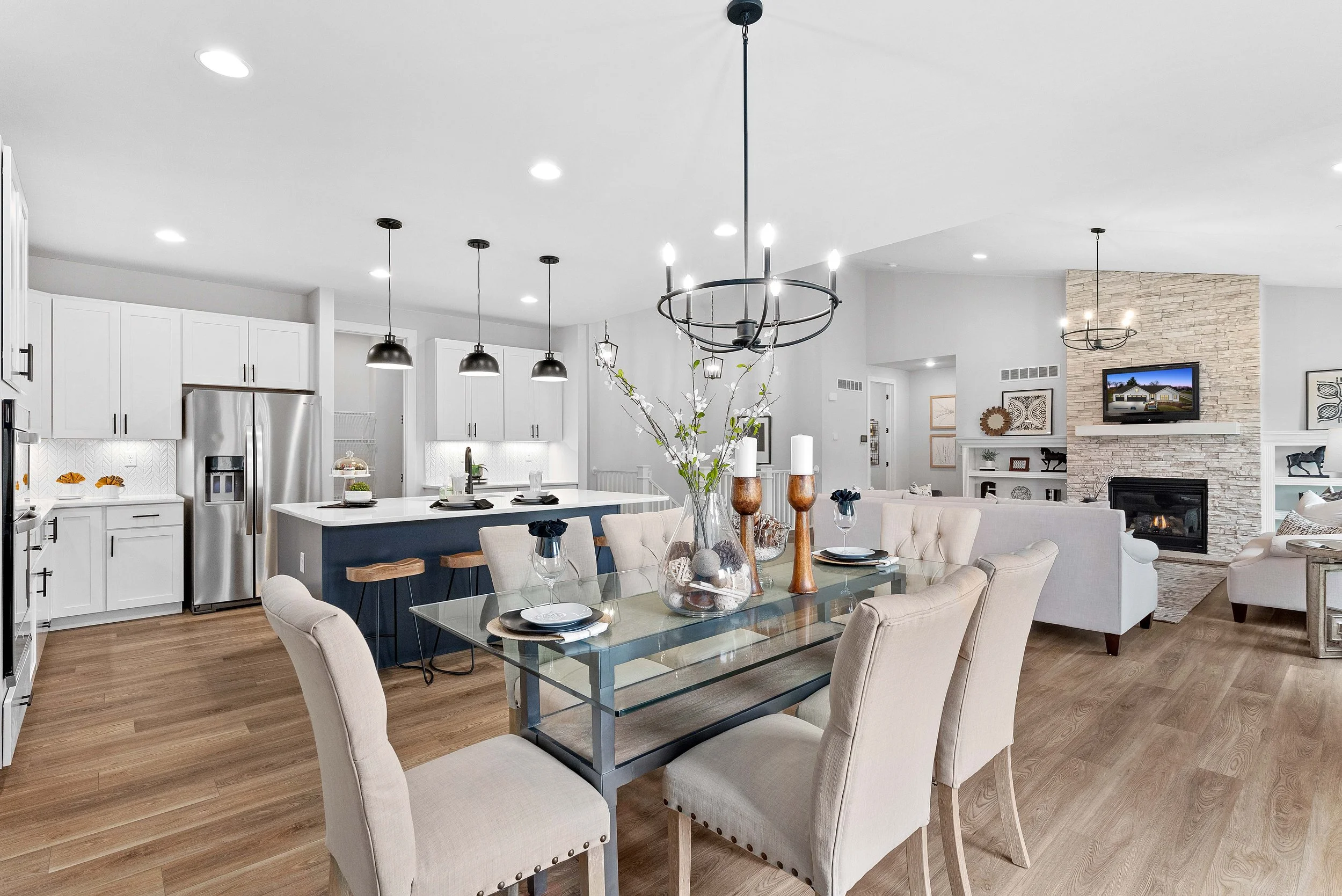 Open-concept living room and kitchen with white cabinetry, stainless steel appliances, a modern chandelier over a dining table, and a stone fireplace with a mounted TV.