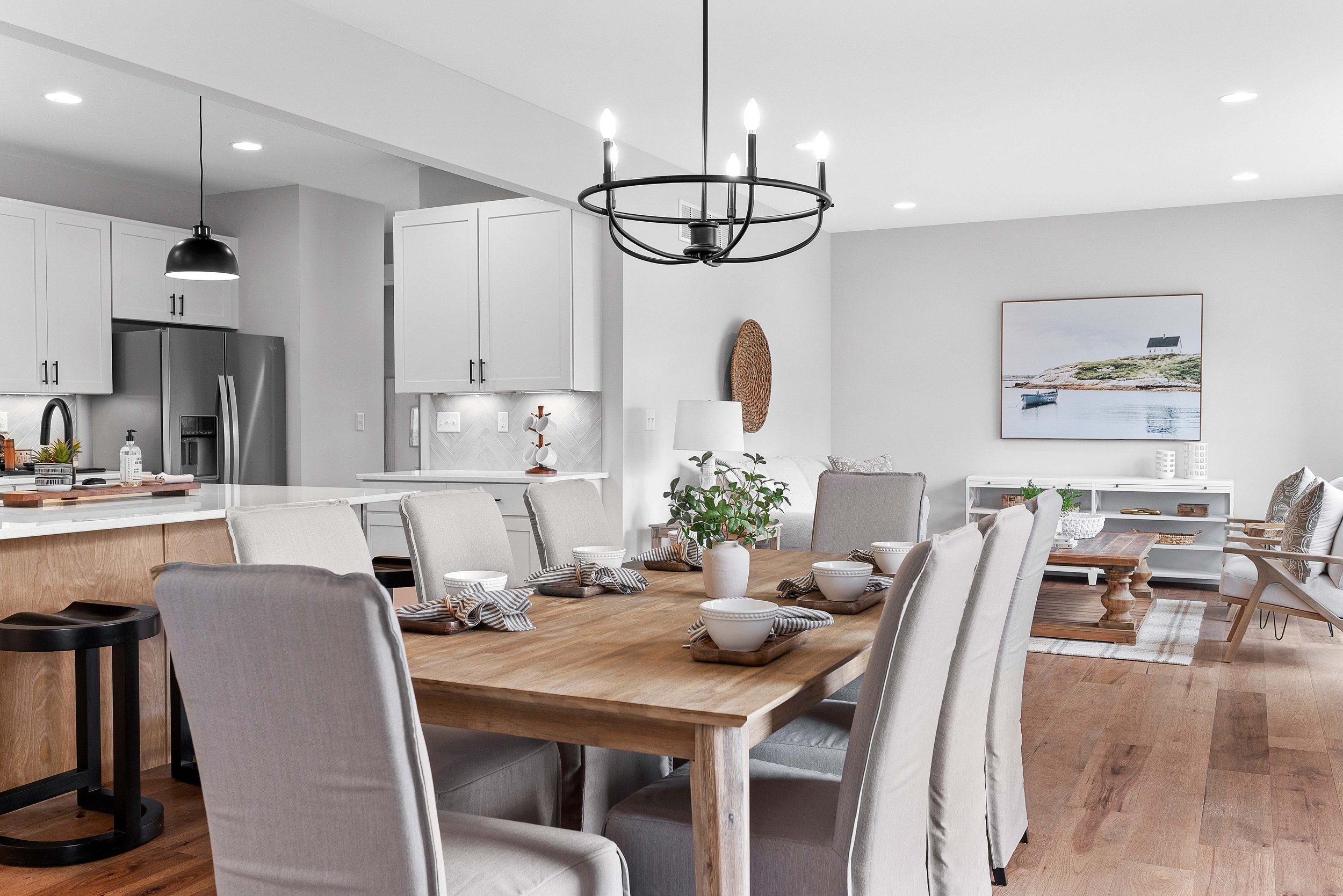 Modern open-concept living and dining area with white walls, wooden flooring, a wooden dining table with eight upholstered chairs, a chandelier, a white sideboard, a lounge area with a coffee table, a painting of a coastal scene, and kitchen applianc