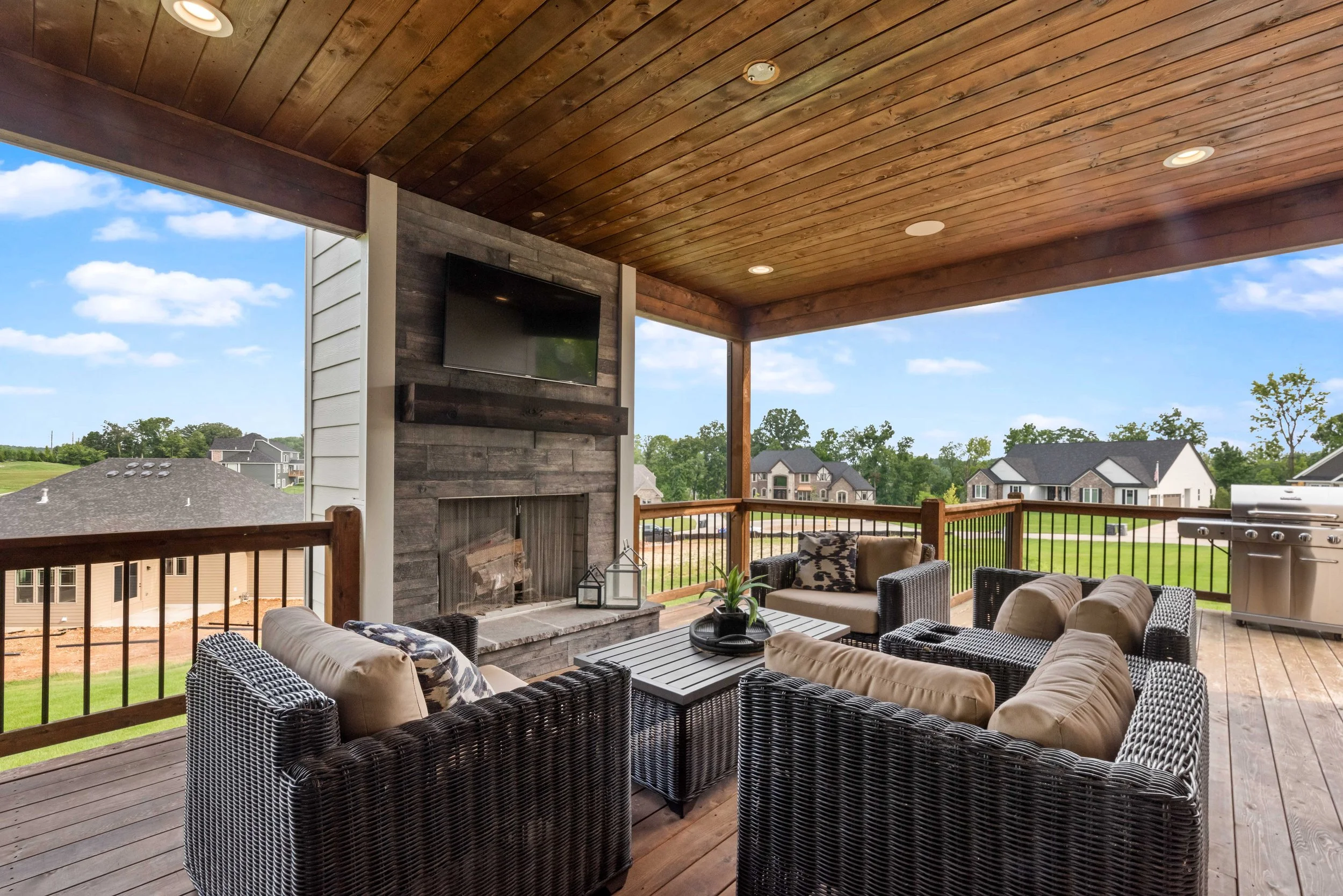 Covered outdoor patio with wicker chairs and beige cushions, a fire fireplace, a wall-mounted TV, a wooden ceiling, and a grill, overlooking a backyard with grass and nearby houses.