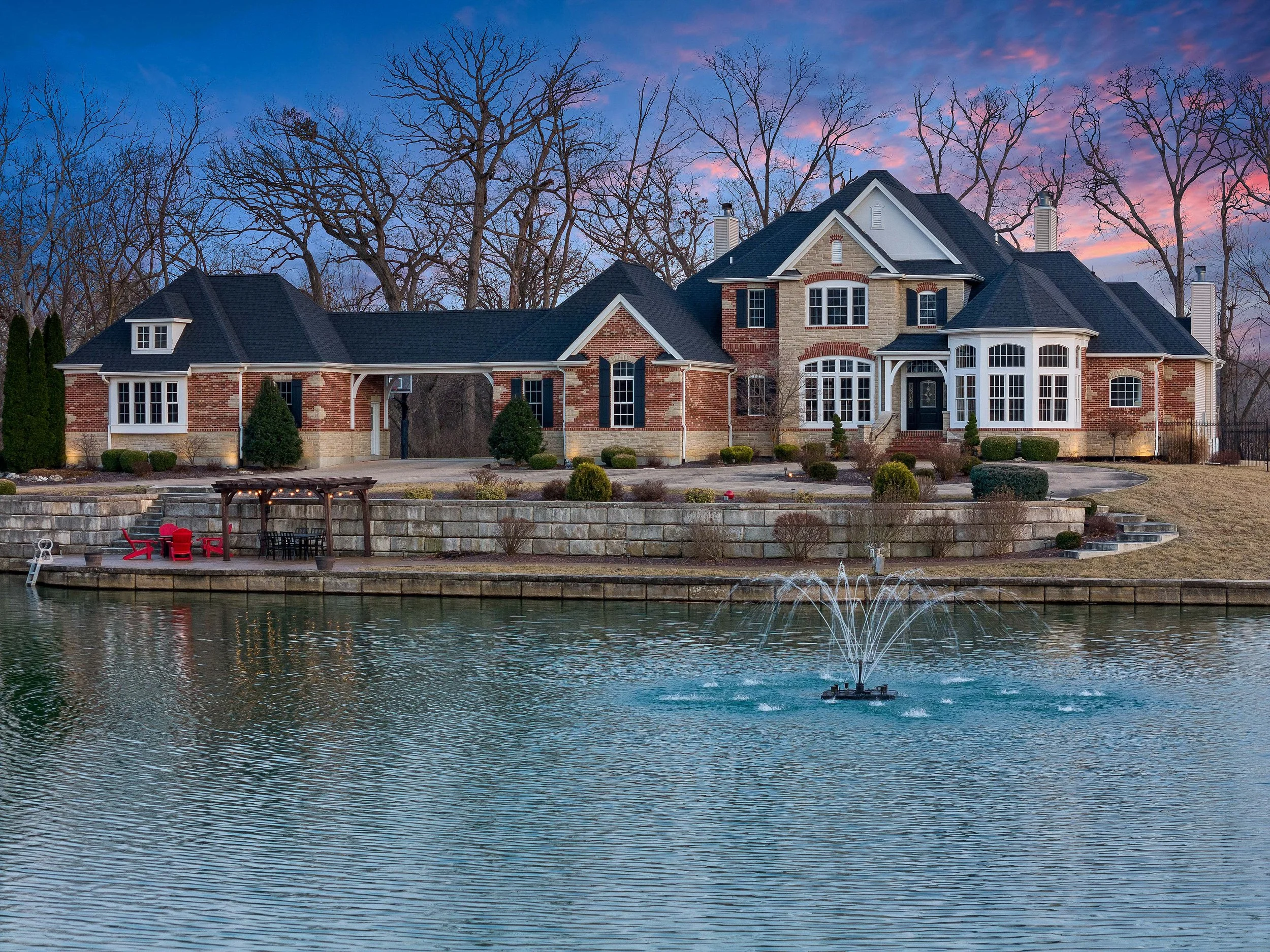A large brick house with a black roof situated beside a water body with a fountain and outdoor seating area, during dusk with a colorful sky and leafless trees in the background.