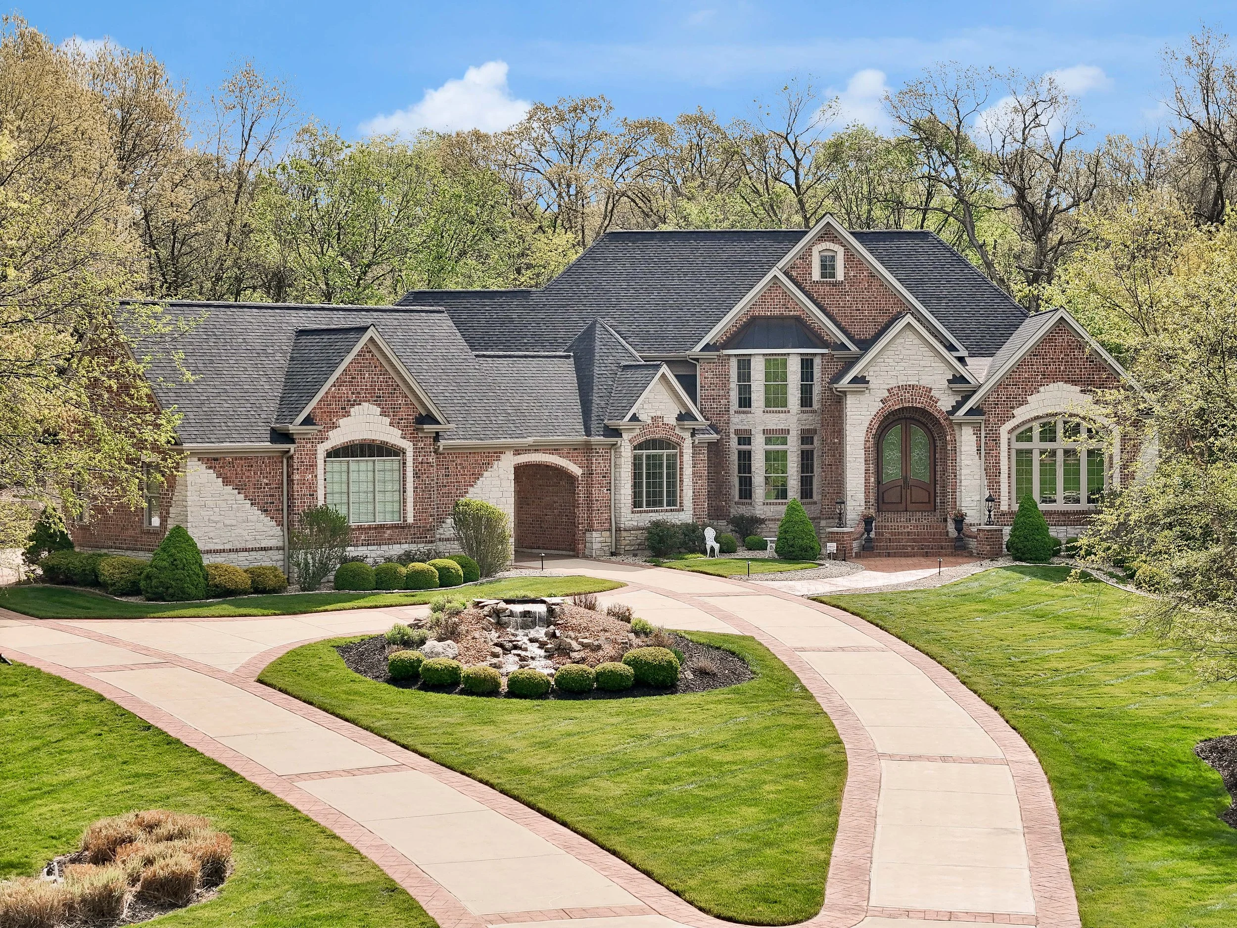 Large brick and stone house with a landscaped front yard, curved walkway, and small waterfall feature. Trees in the background, clear sky, and well-maintained garden.