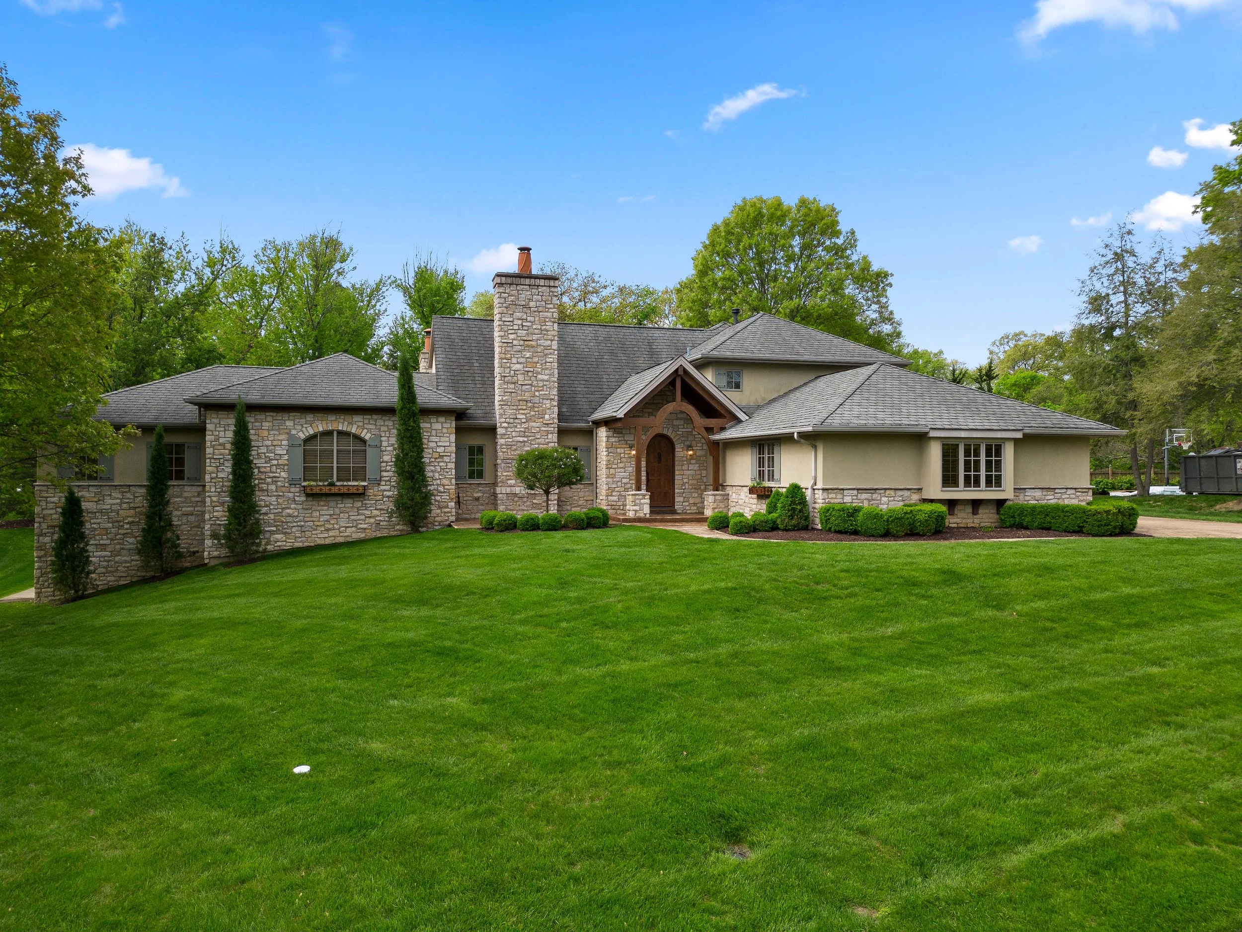 A large house with stone and beige siding exterior, surrounded by well-manicured green lawn and trees, under a partly cloudy blue sky.