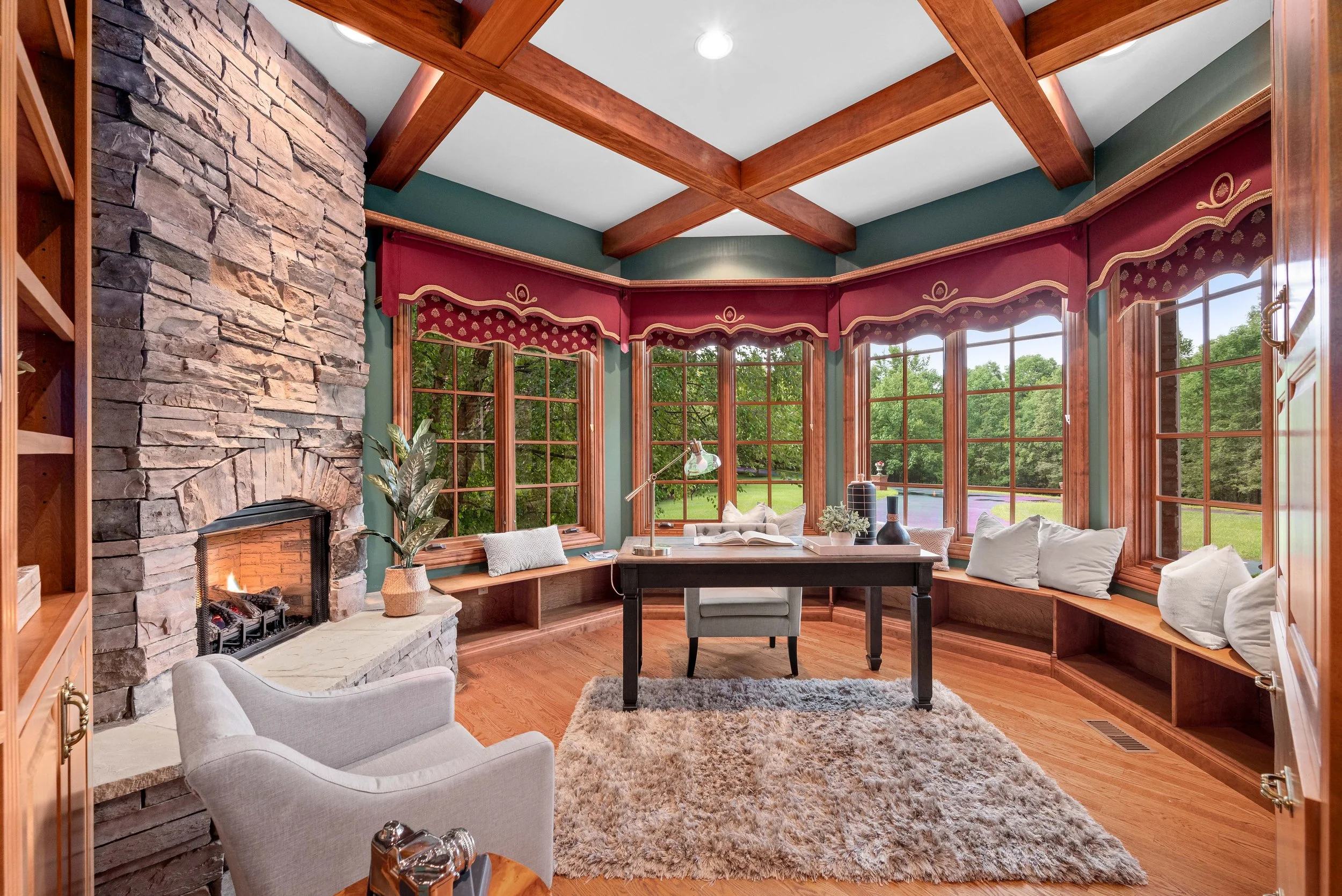 A cozy reading nook with a stone fireplace, bay windows with red valances, a wooden desk in front of the windows, a white armchair, and a plush rug.