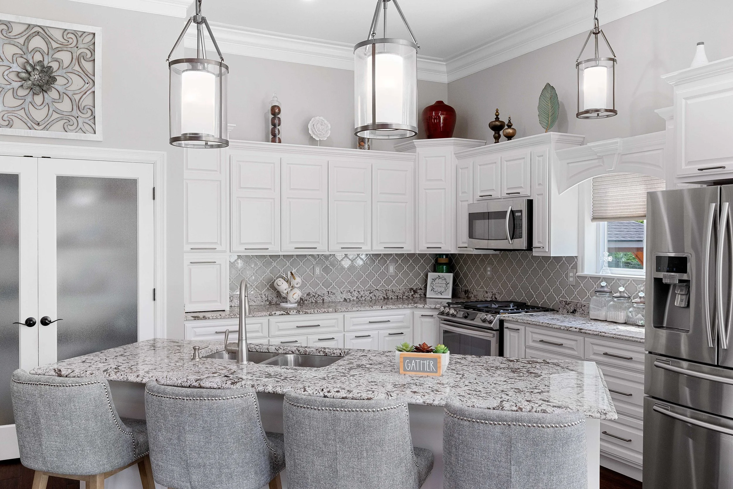 Modern kitchen with white cabinetry, granite countertops, stainless steel appliances, and gray upholstered bar stools around an island. Decor includes wall art, vases, and a window with blinds.