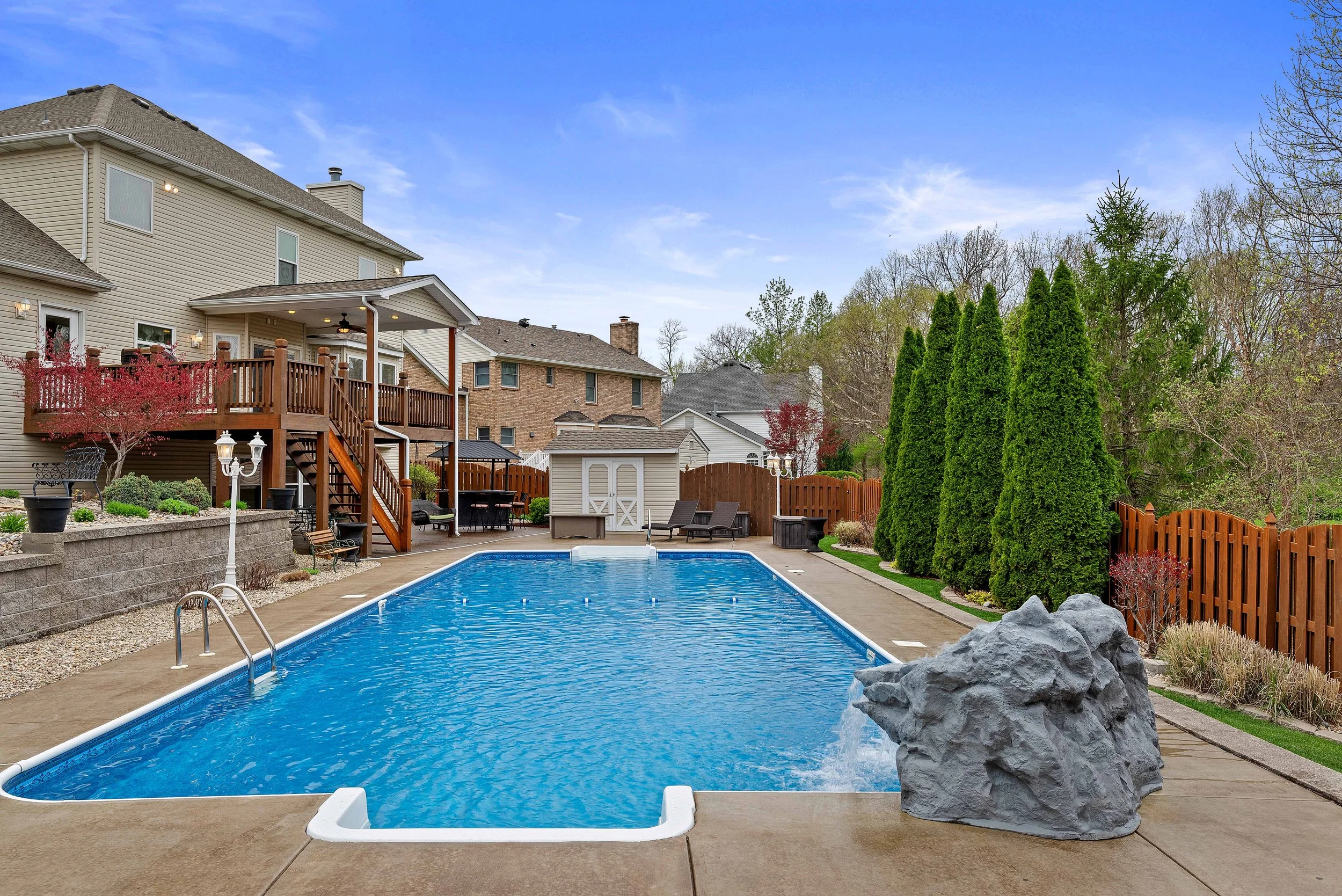 Backyard with a swimming pool, wooden deck, outdoor furniture, trees, and a wooden fence.